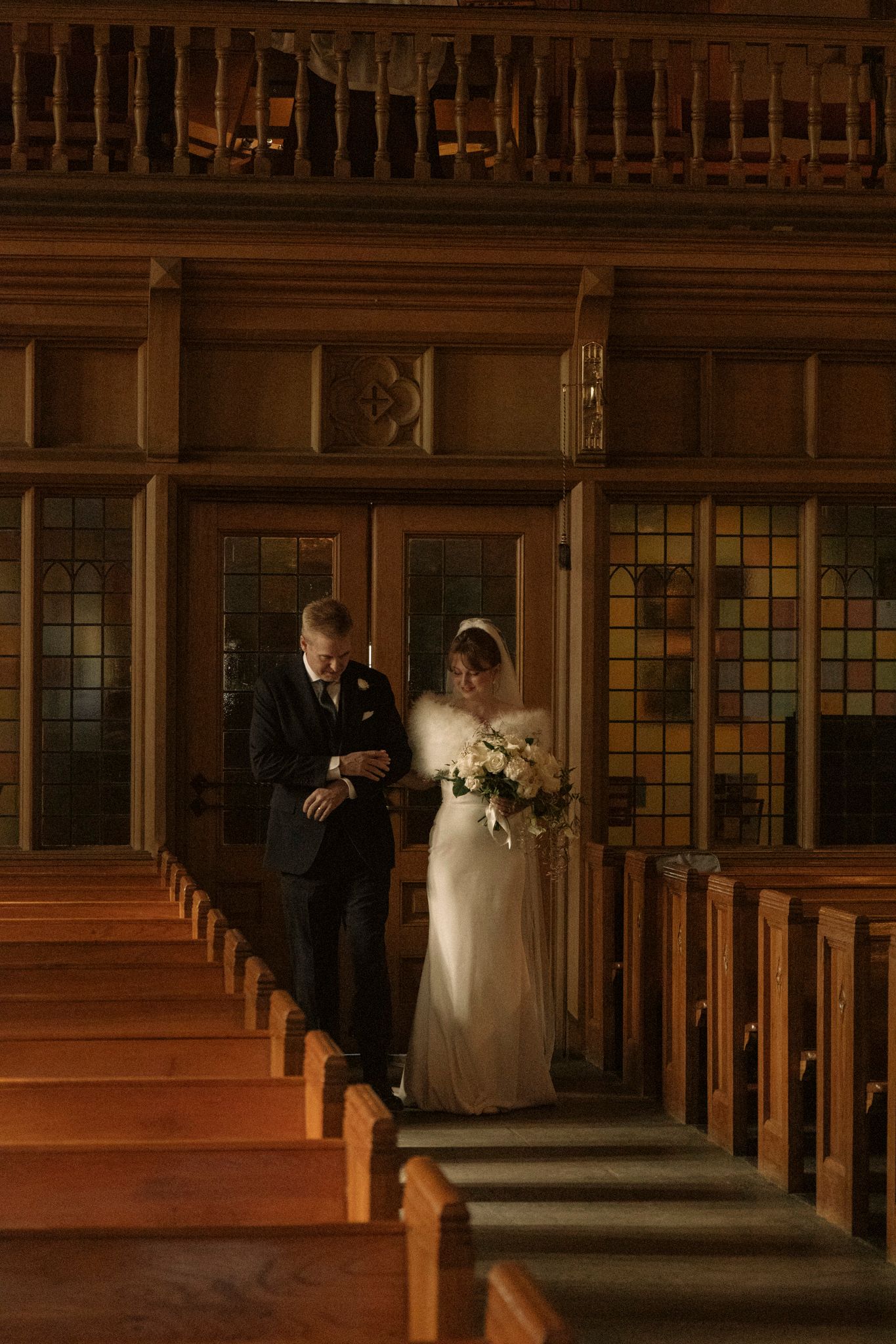 Bride in white gown walks down church aisle with escort past wooden pews and stained glass windows.