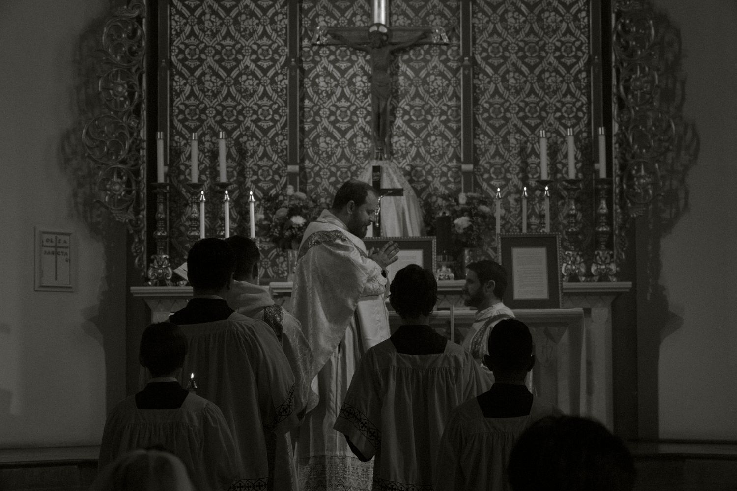 Black and white photo of priest and altar servers celebrating Mass at ornate church altar.
