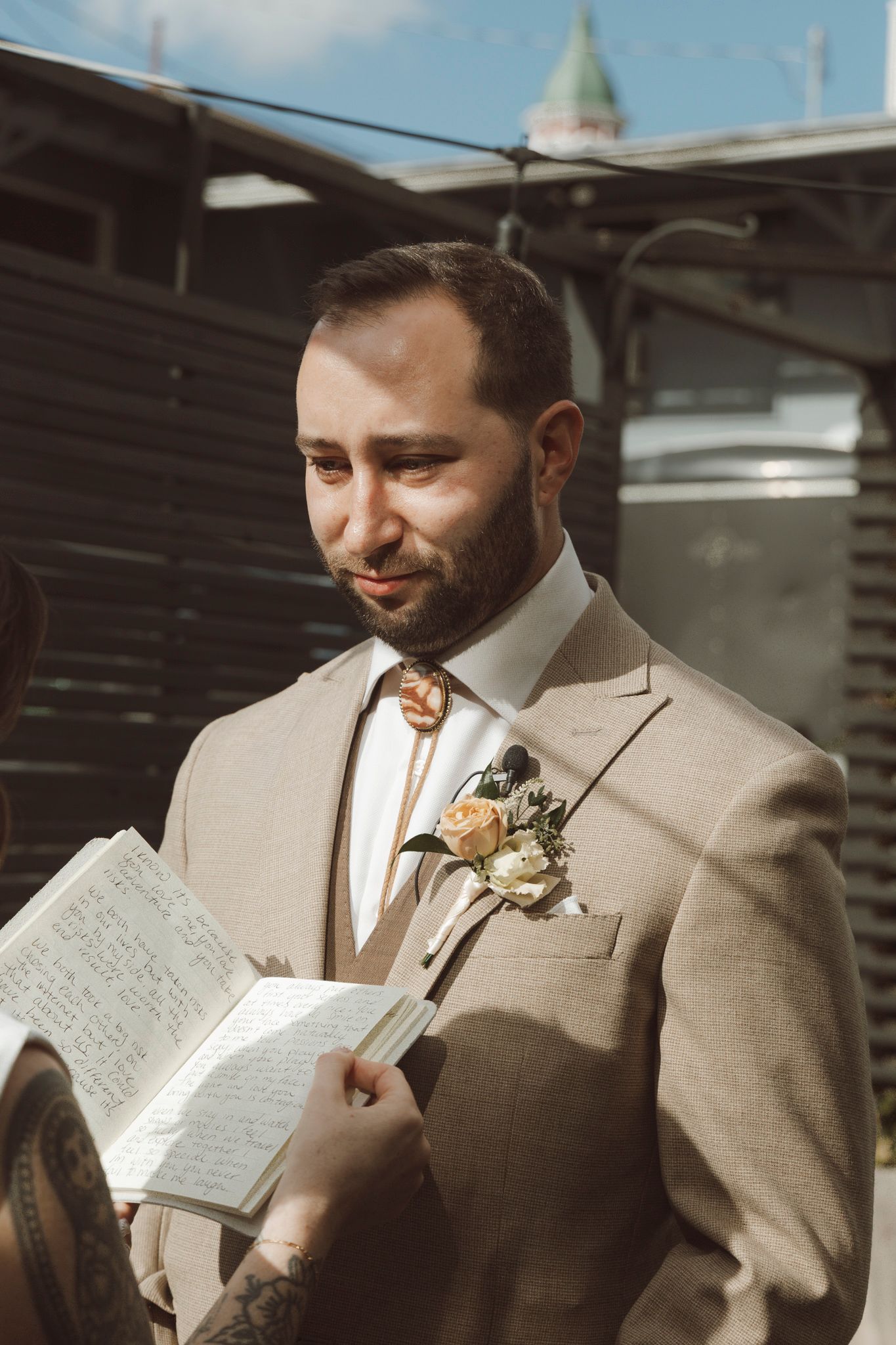 A groom in a beige suit with boutonniere reads from a small book during an outdoor ceremony.