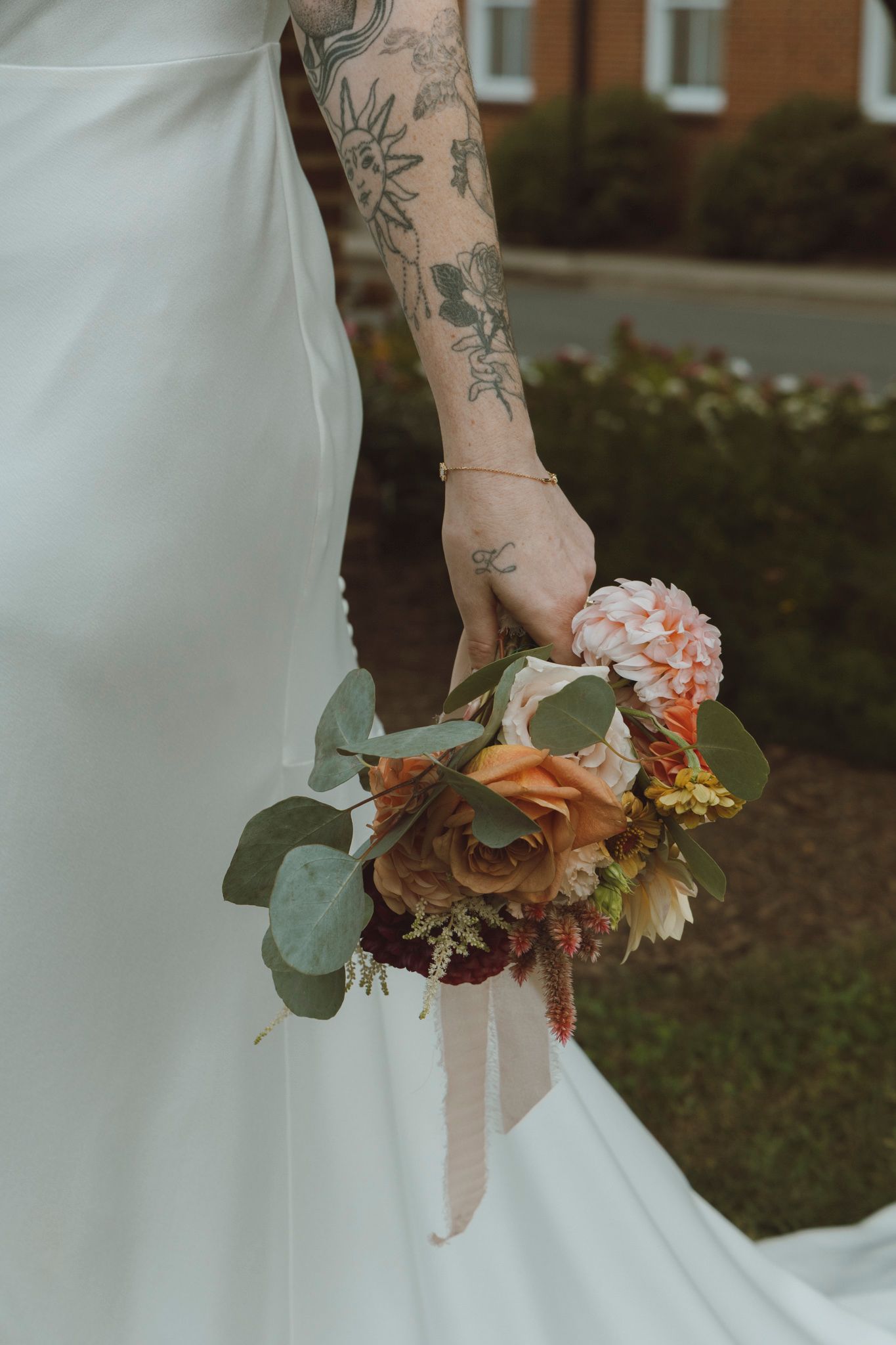 Close-up of tattooed arm holding romantic peach and eucalyptus wedding bouquet against white dress.
