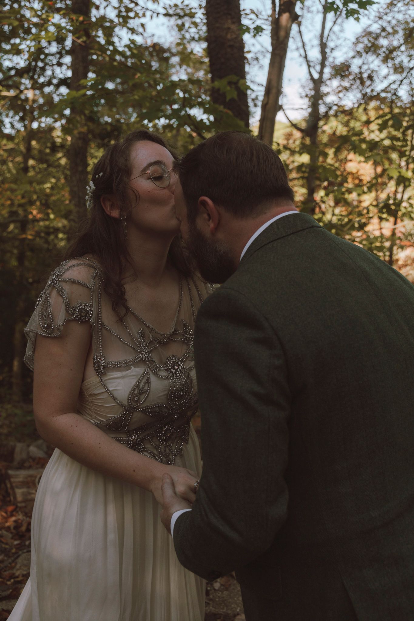 A romantic couple shares tender moments in a wooded area during sunset, wearing formal wedding attire.