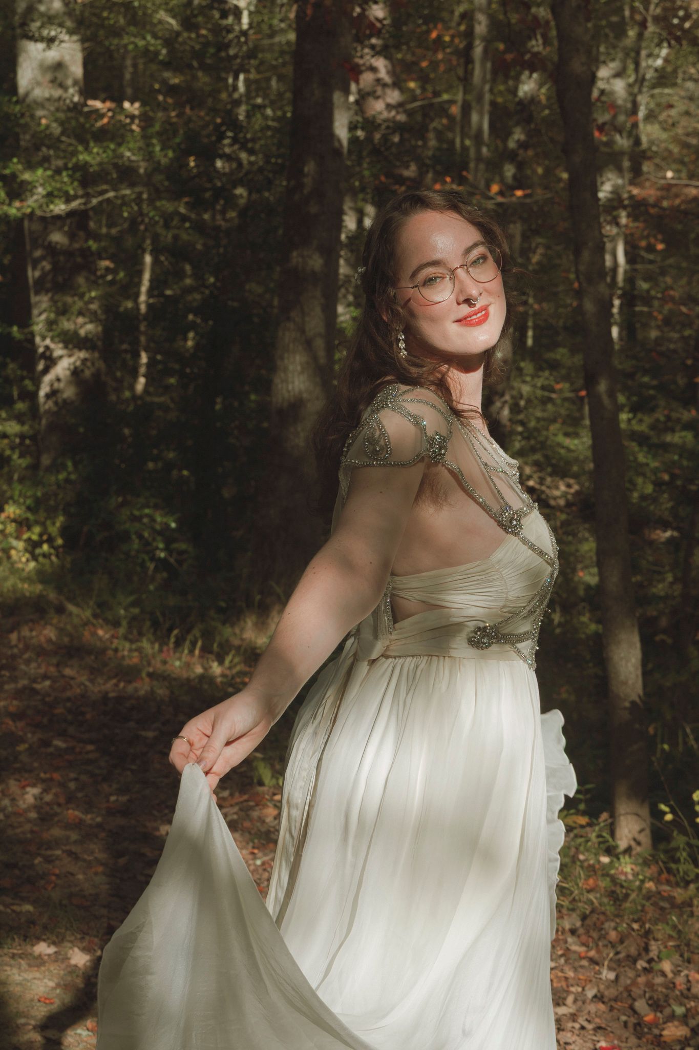 A person in a flowing white dress and glasses poses gracefully in a sunlit forest setting.