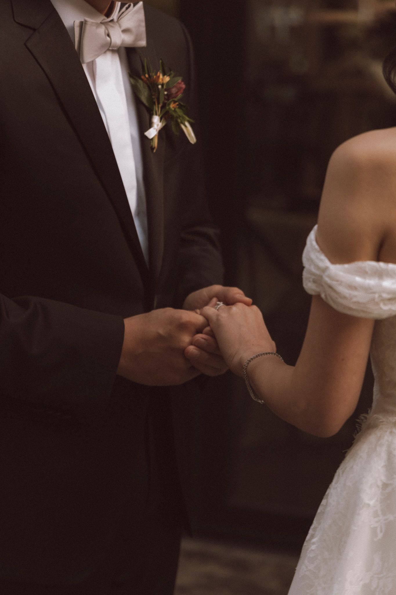 Close-up of bride and groom holding hands during wedding ceremony showing white dress and black tuxedo details.