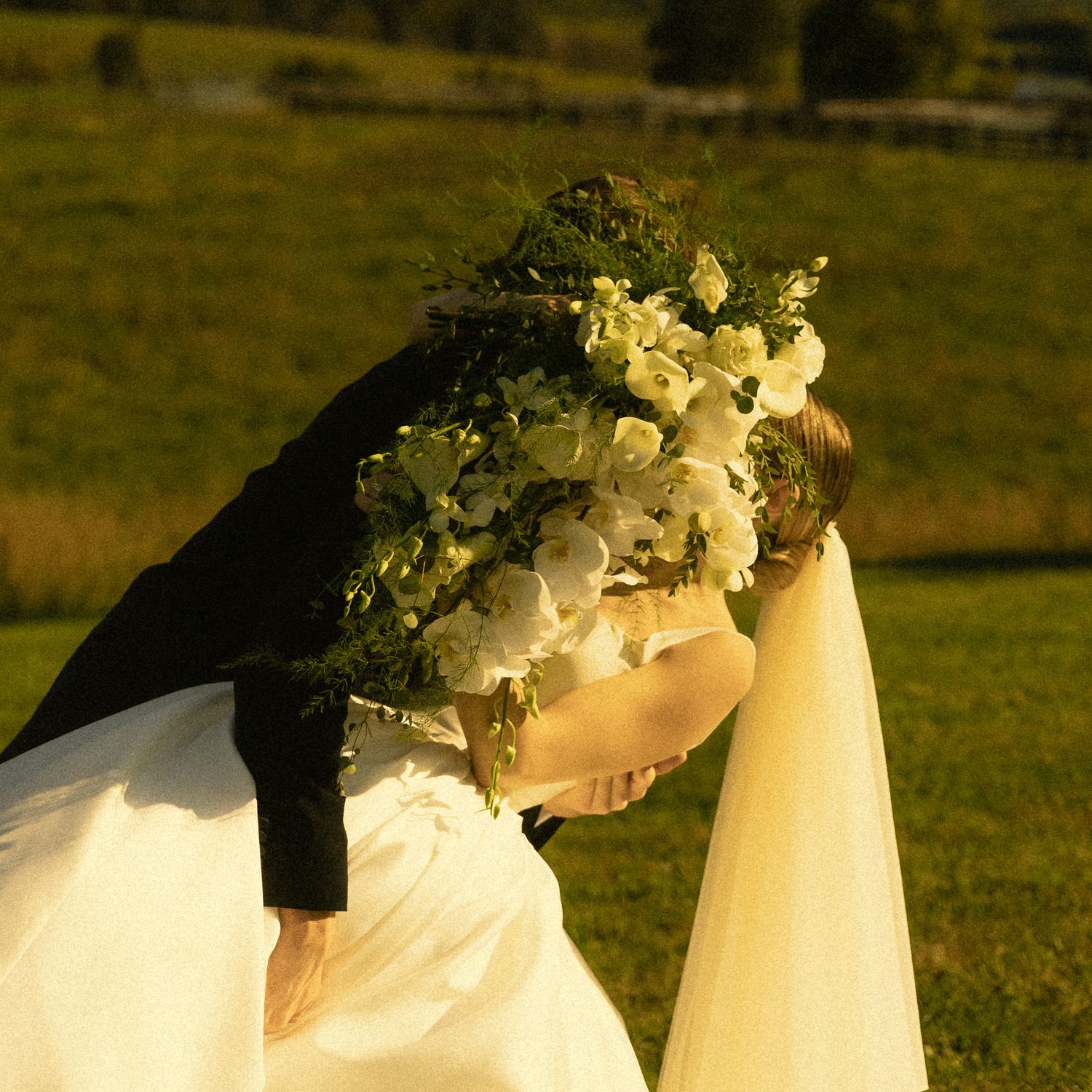 Bride in a white gown holding a lush white floral bouquet embraces a groom in a dark suit on a sunlit grassy hillside.