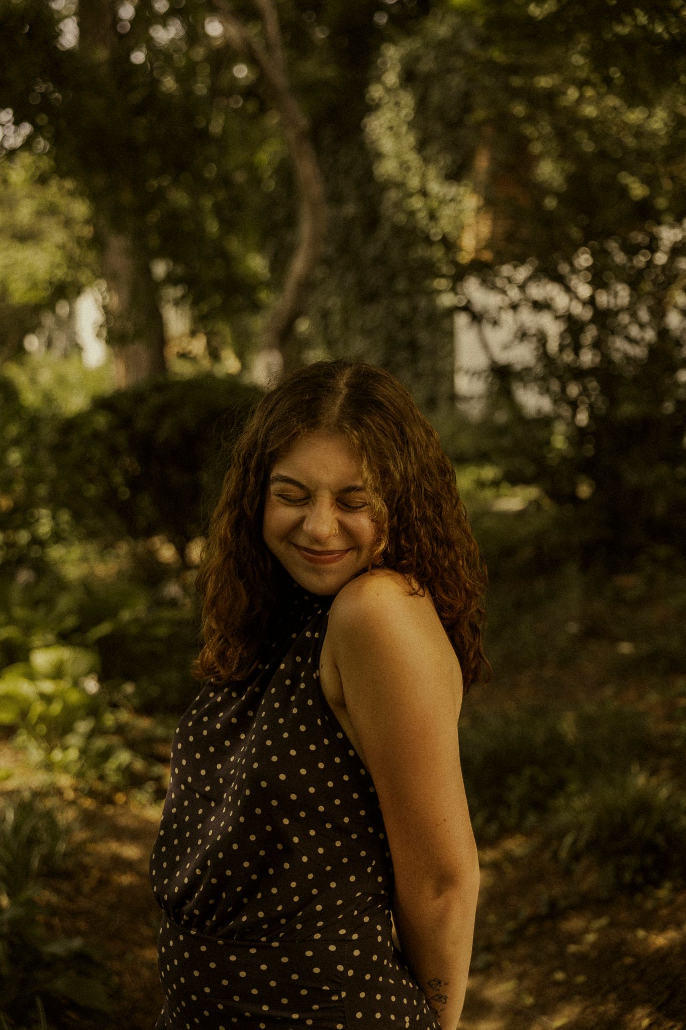 A young woman with curly hair smiles warmly in a sunlit garden, wearing a black polka dot sleeveless dress.