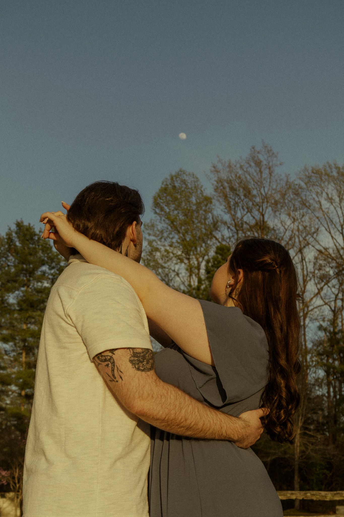 A couple embraces outdoors at dusk with a full moon visible in the clear blue sky above the trees.