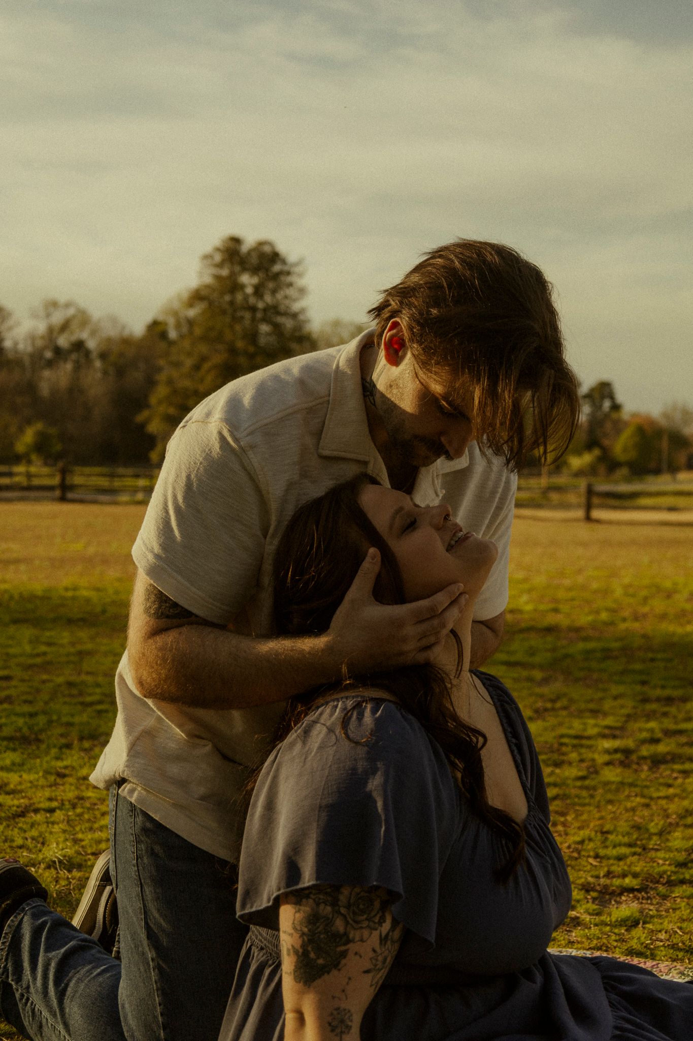 A couple shares a tender moment at golden hour in a park, with the man leaning down to kiss the woman sitting on the grass.