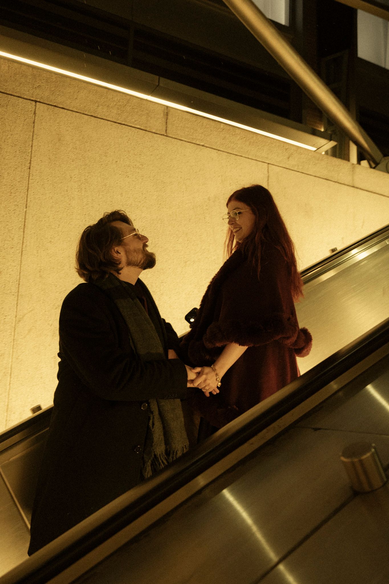 A couple holds hands while riding an escalator in warm lighting conditions.