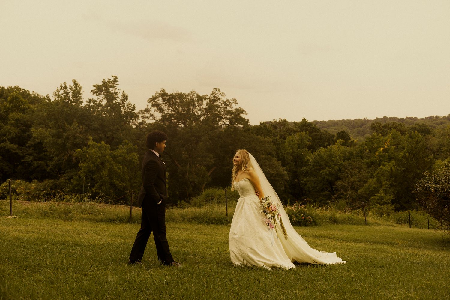 Bride and groom walking through lush green field at dusk during outdoor wedding ceremony.