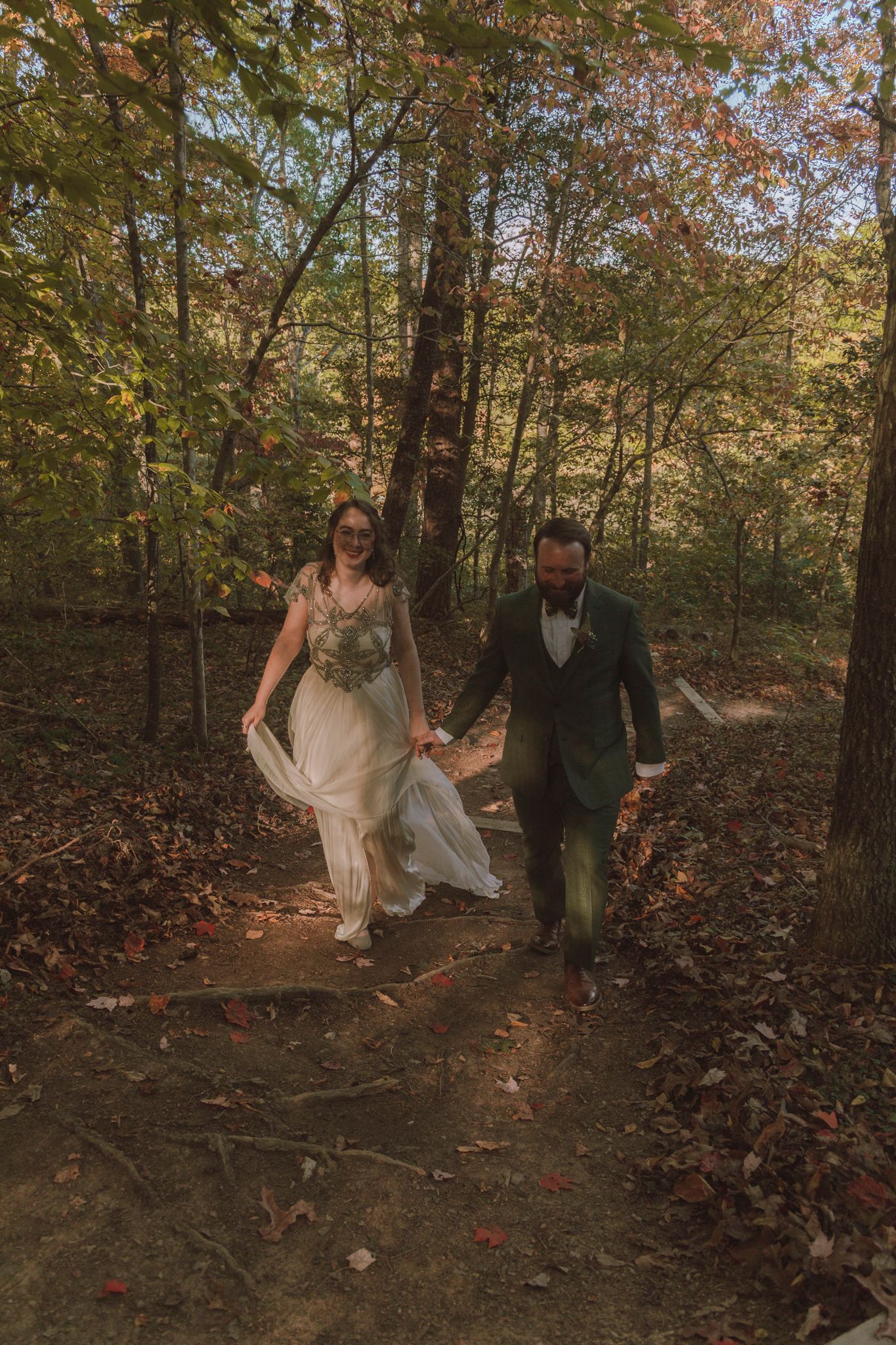 A couple walks together through a scenic autumn forest path with fallen leaves during their wedding celebration.