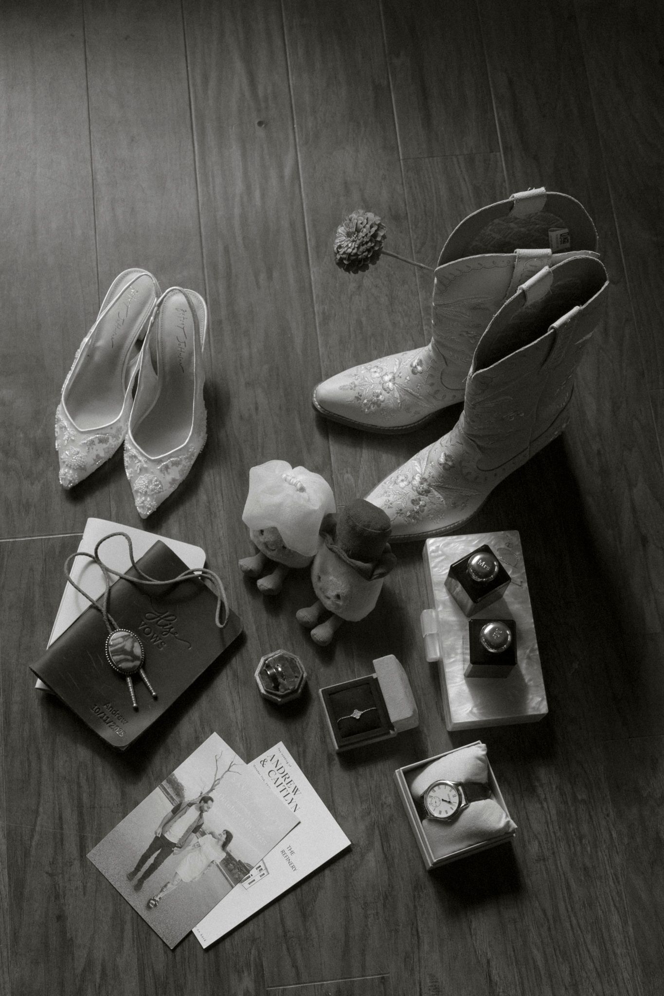 Black and white photo of western boots, lace shoes, and wedding accessories arranged on dark floor.