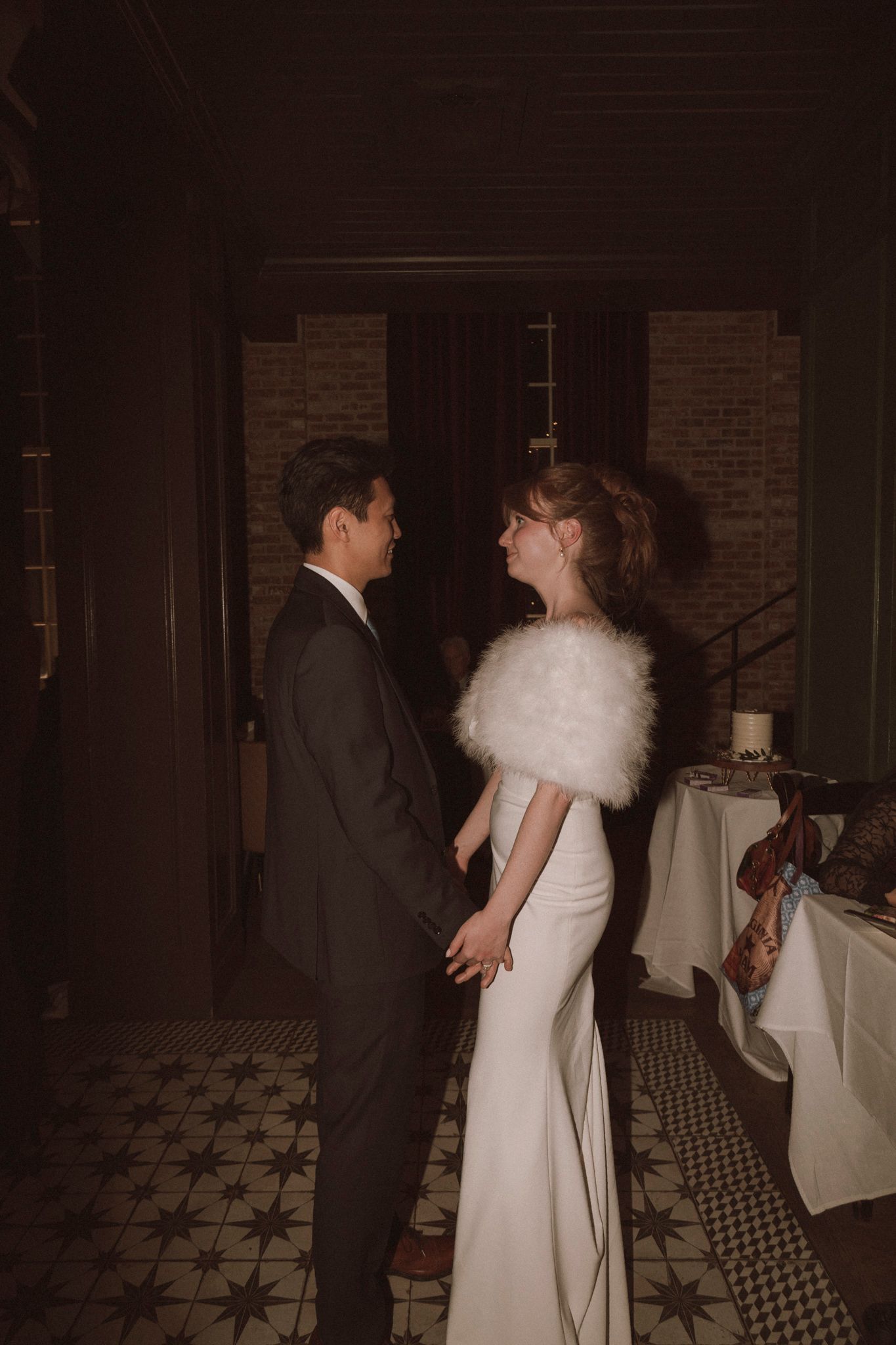 Bride in white gown with fur stole holds hands with groom in dark suit in venue hallway.