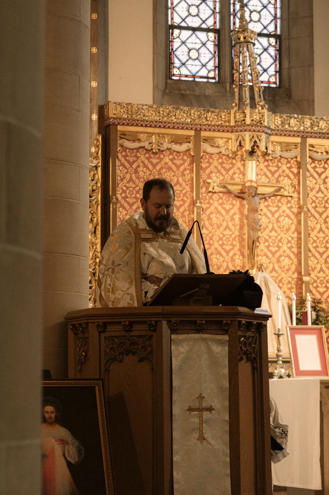 Priest reads from book at ornate wooden pulpit before decorative red and gold church altar.