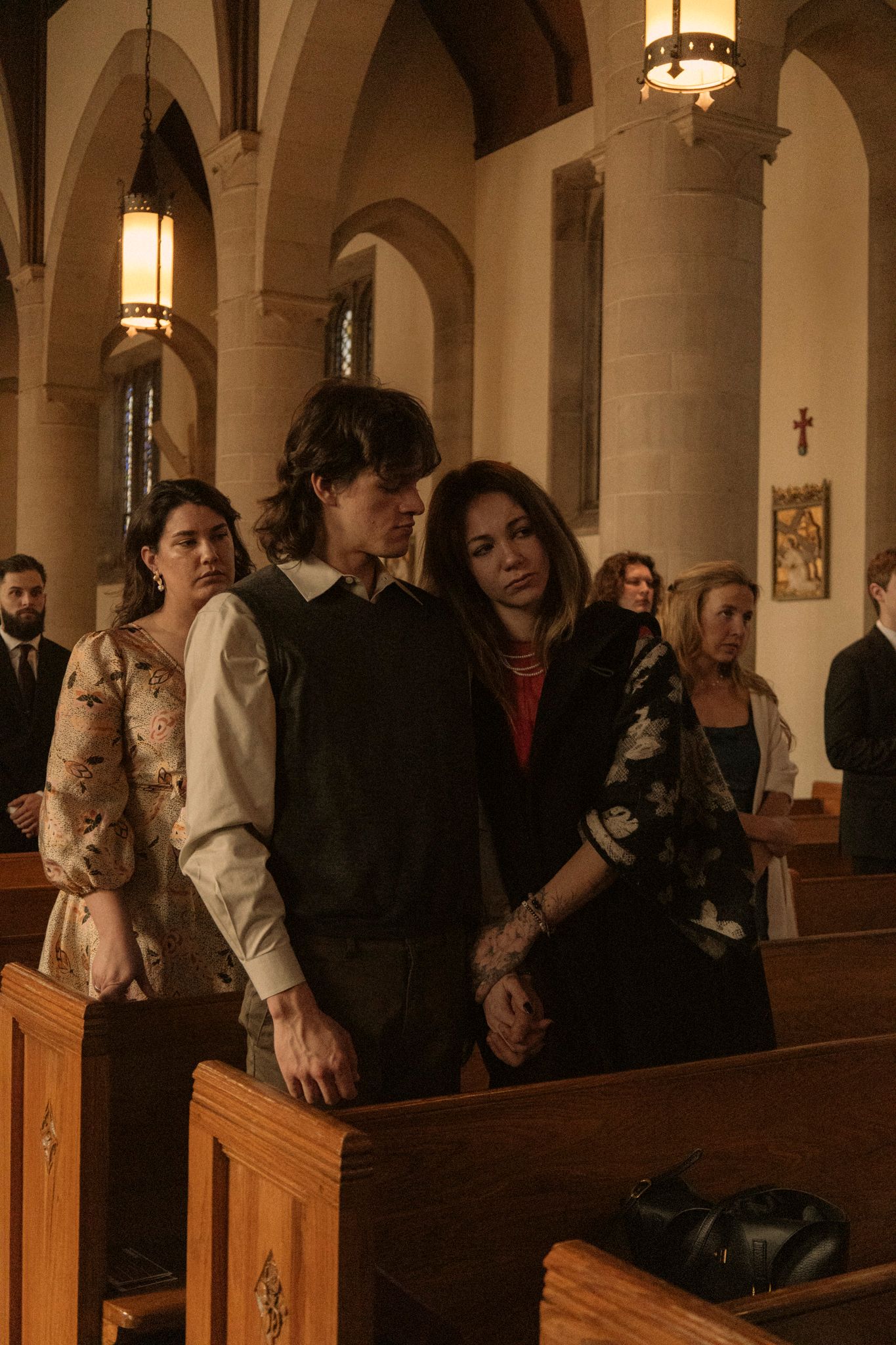 Wedding guests standing in Gothic church pews, including a couple embracing, during a Catholic wedding ceremony.