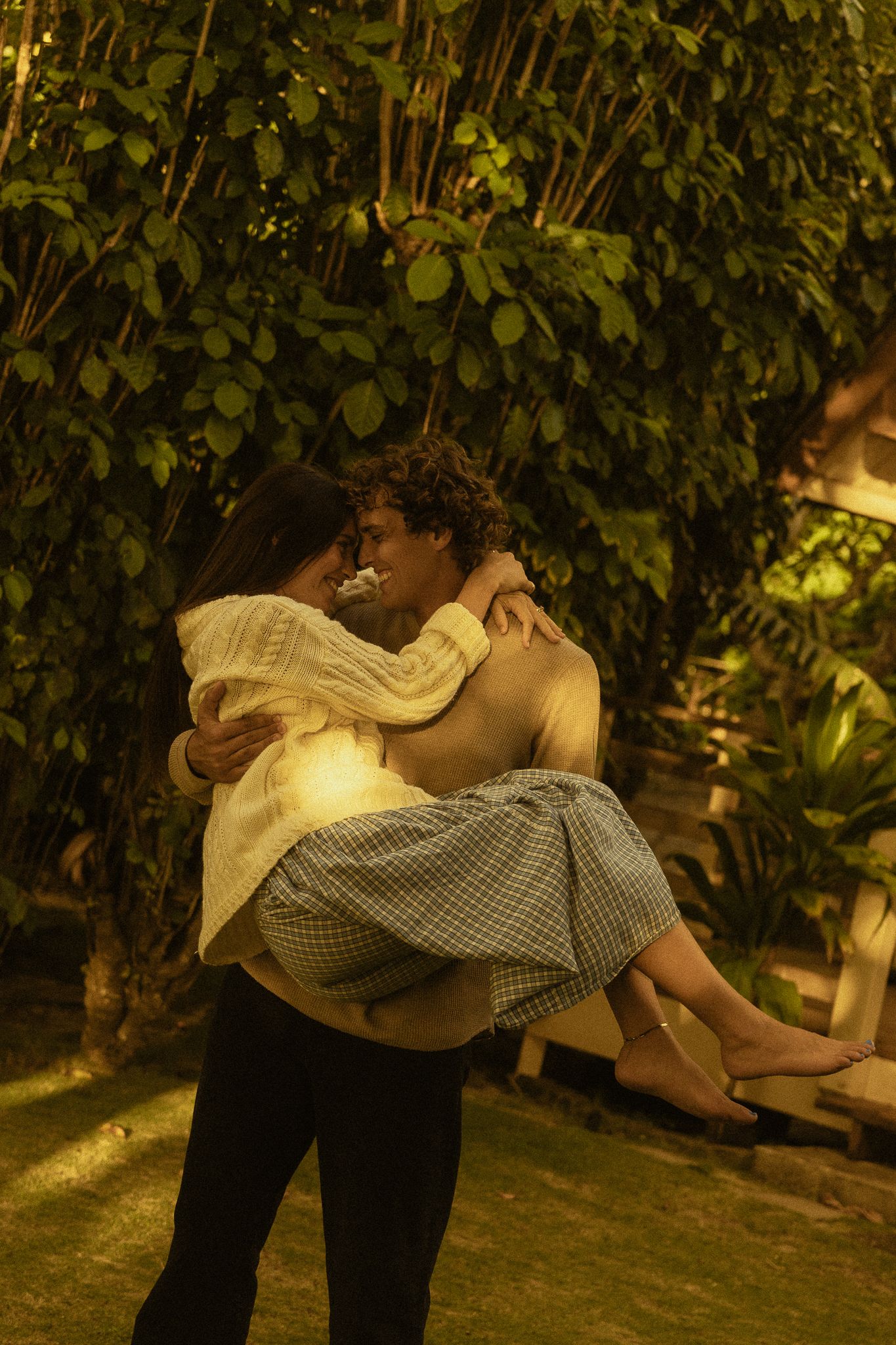 A shirtless man carries a woman in his arms, sharing an intimate moment in a lush green garden setting.