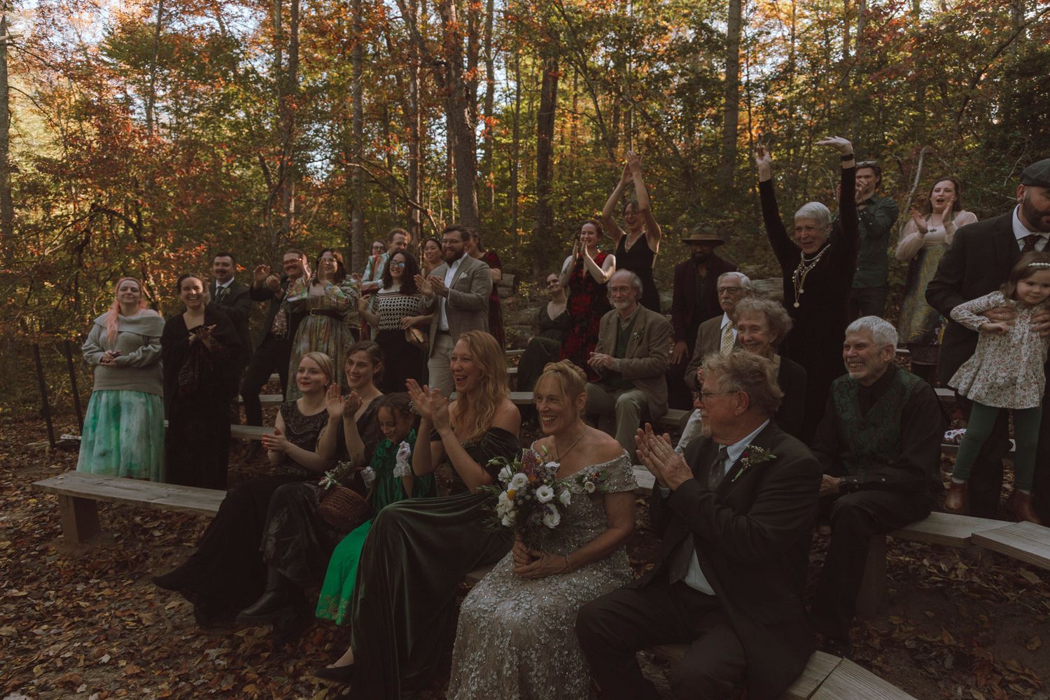 A group of wedding guests gather in a wooded outdoor ceremony area during autumn with golden sunlight filtering through trees.