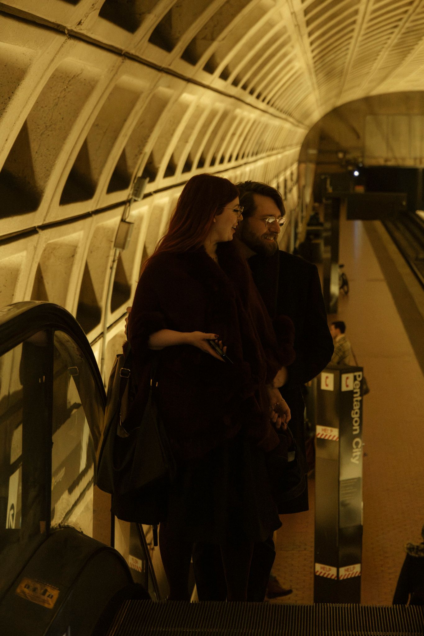 People standing on an illuminated escalator in a modern subway station with arched architectural details.