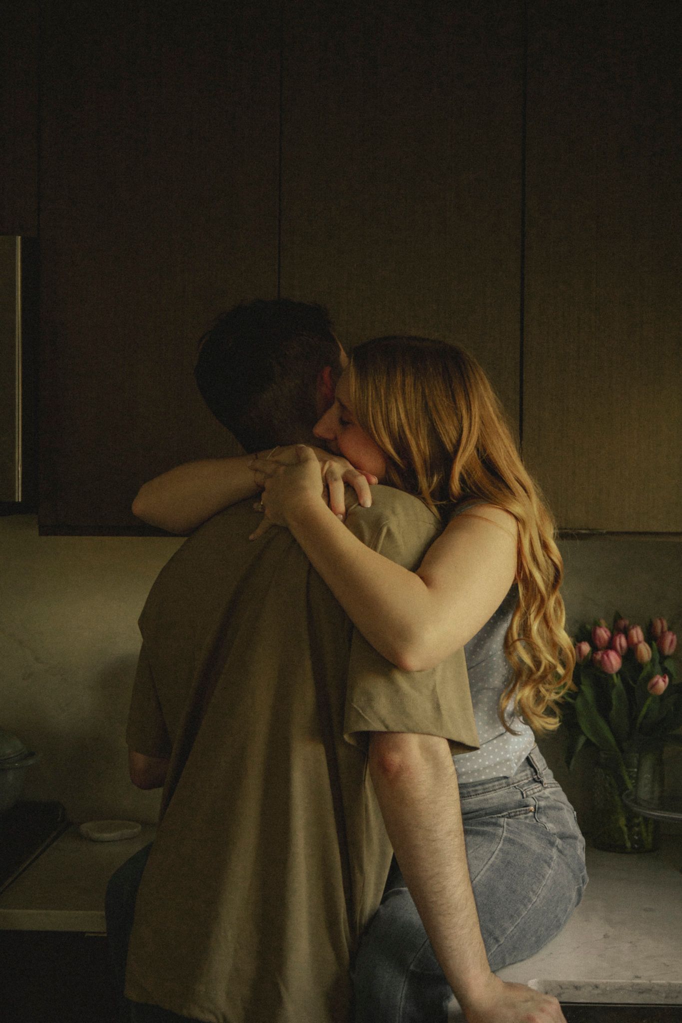 A couple shares a passionate kiss in a dimly lit kitchen, with pink tulips visible in the background.