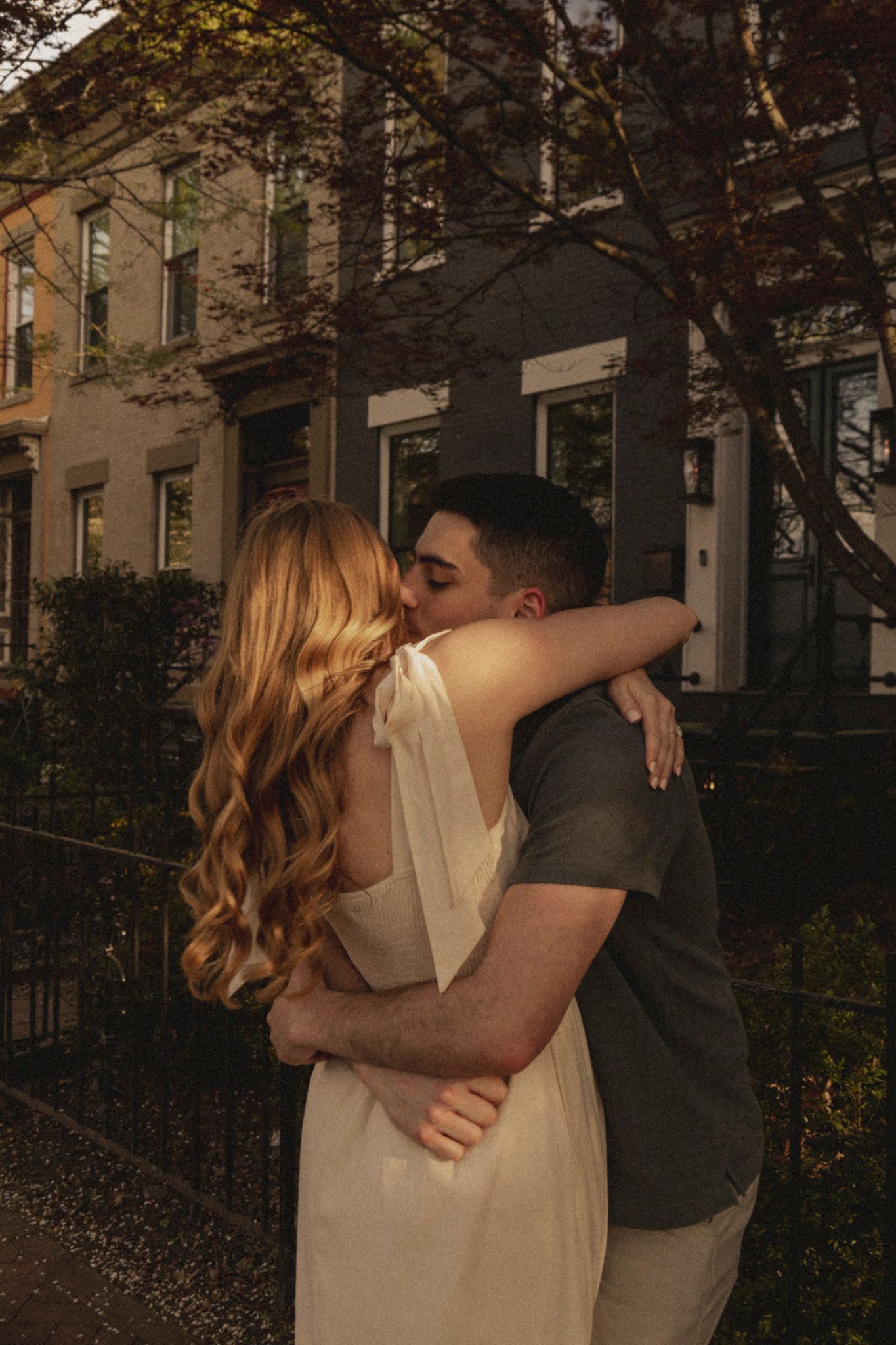A couple shares a romantic kiss while embracing outdoors near brownstone buildings with autumn trees.