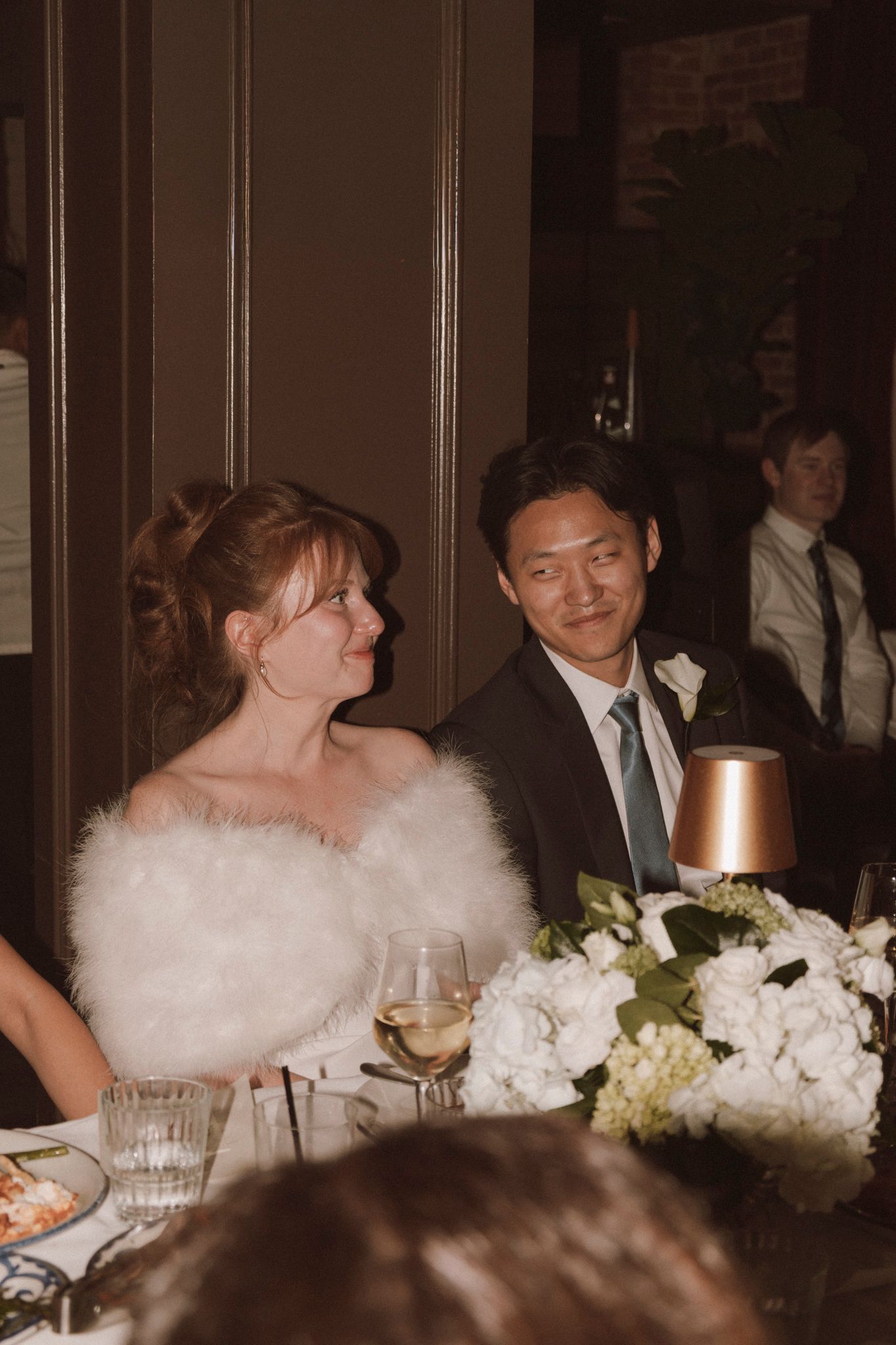 Bride in white feather wrap and groom in suit smiling together at wedding reception dinner table.