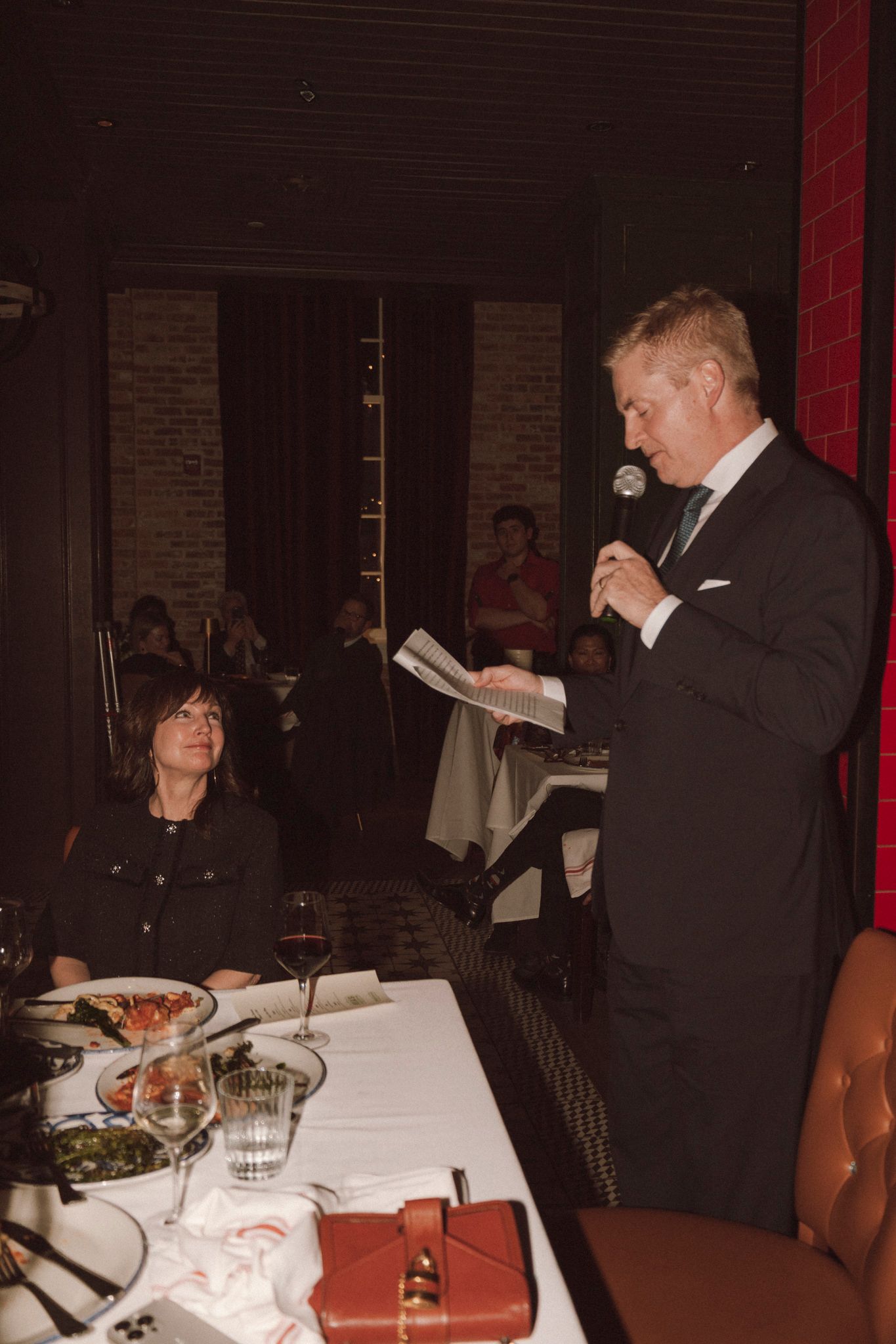 Man in black suit delivers speech at microphone during formal wedding reception dinner.