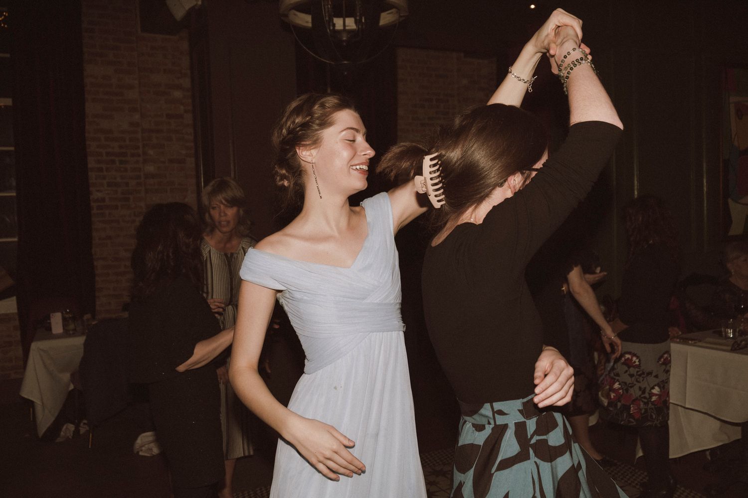 Two women dancing together at a wedding reception in a dimly lit venue, one wearing a white off-shoulder gown.