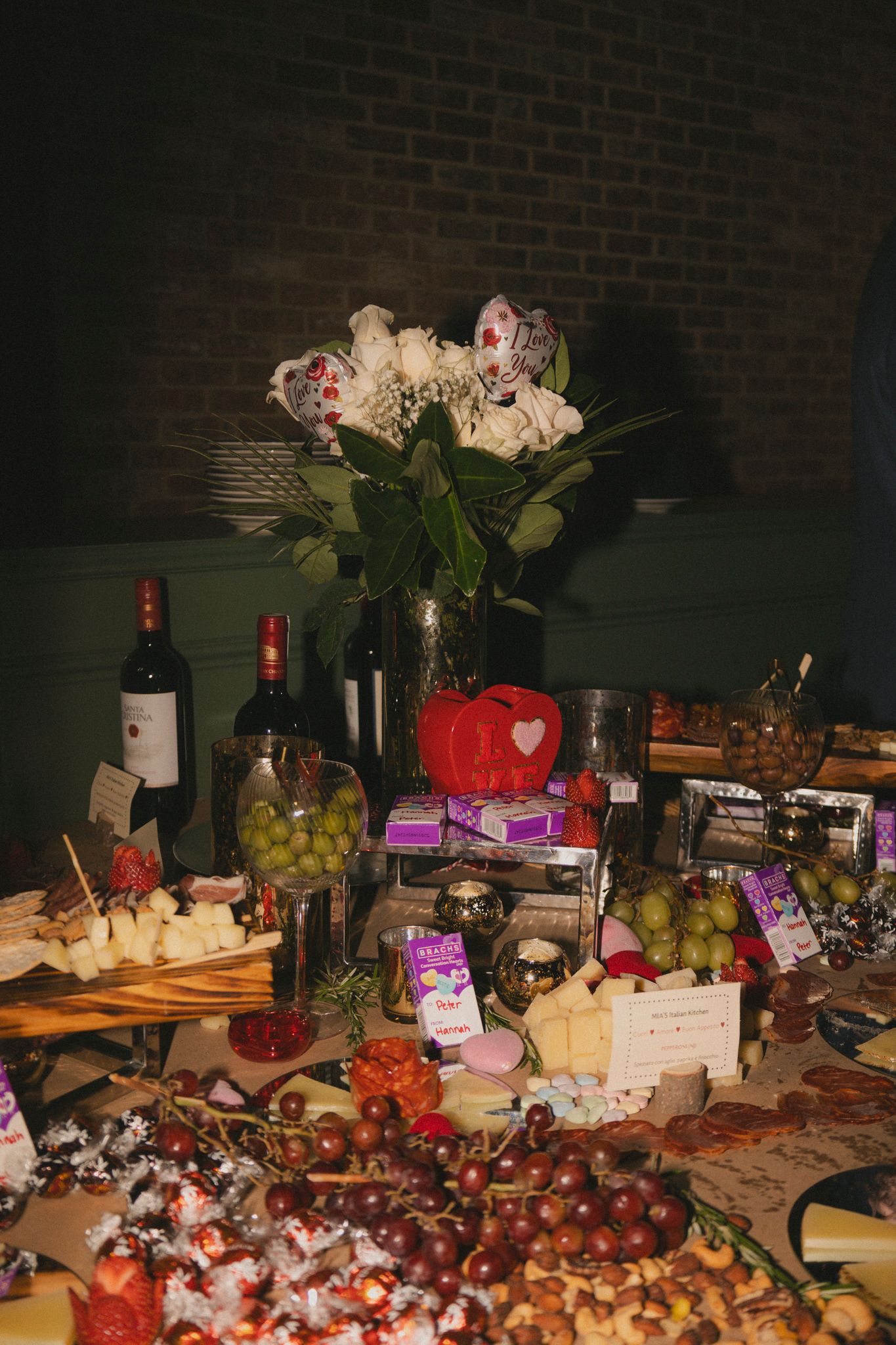 Elegant party grazing table with wine bottles, grapes, cheese, chocolates, and a floral centerpiece with white roses.