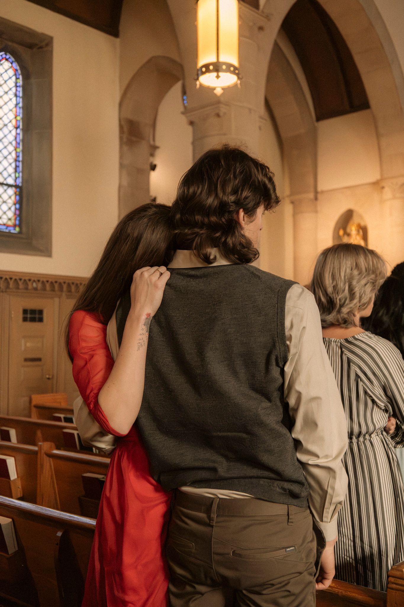 Woman in red dress leaning head on mans shoulder inside church during wedding ceremony.