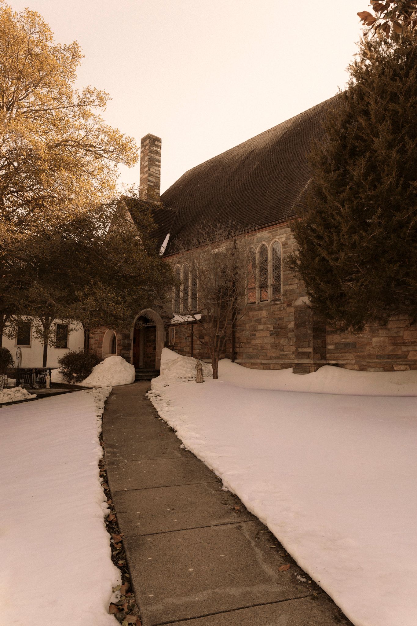 Snow-covered stone church with pathway leading to arched entrance, surrounded by trees in winter landscape.