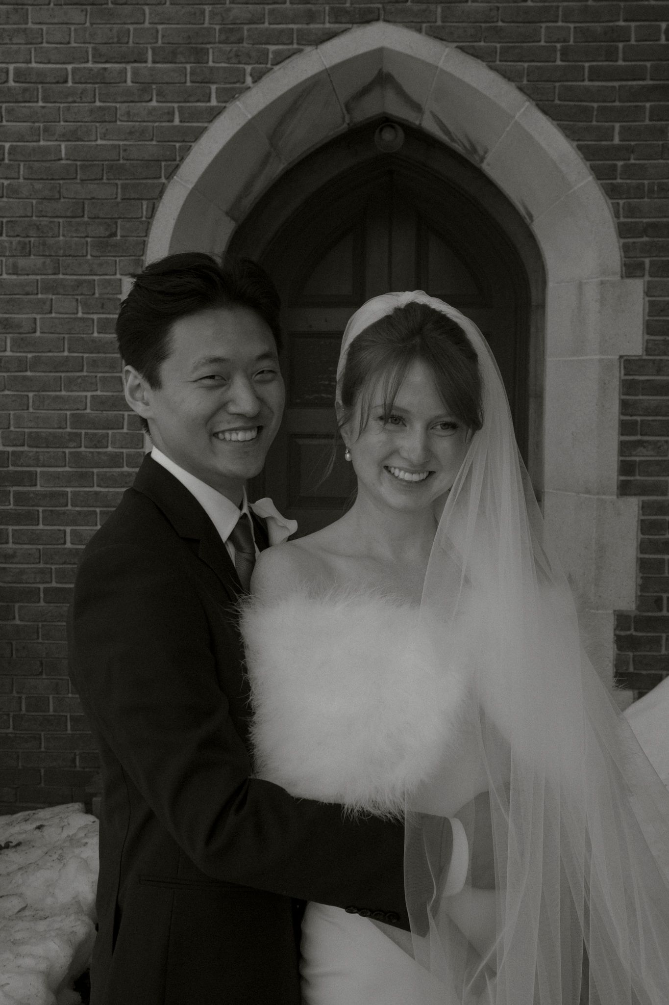 Smiling newlywed couple poses outside a brick church, bride in white gown with veil, groom in dark suit, black and white photo.