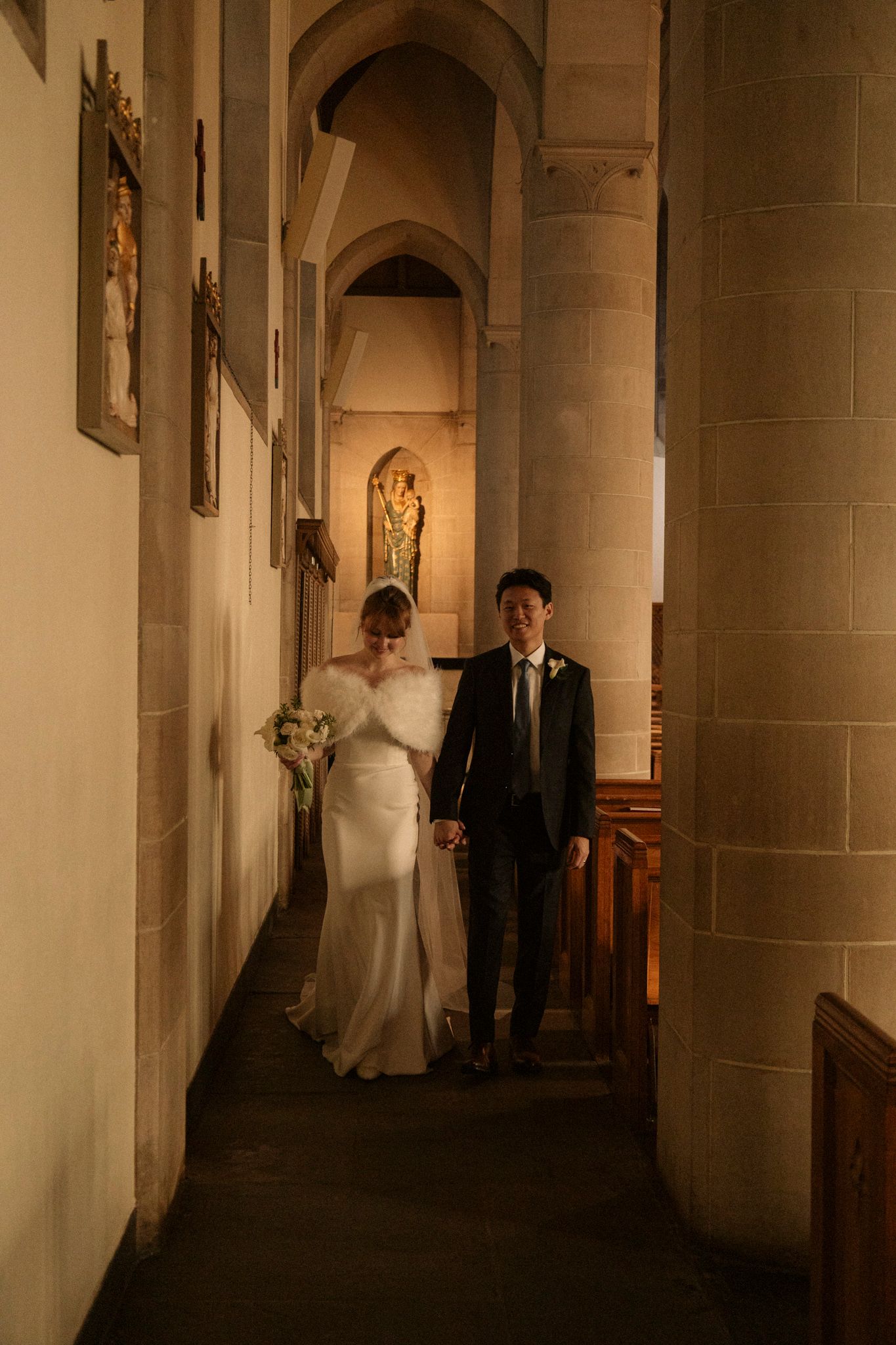 Bride and groom walking hand in hand down a warmly lit stone church aisle during their wedding ceremony.