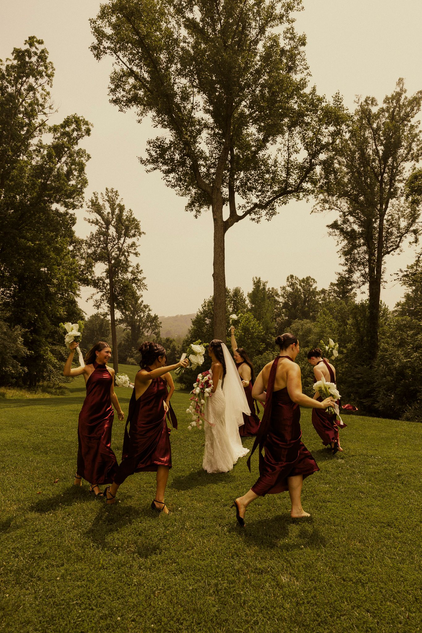 Bridesmaids in burgundy dresses walk with bride in white gown across lush green lawn surrounded by tall trees.