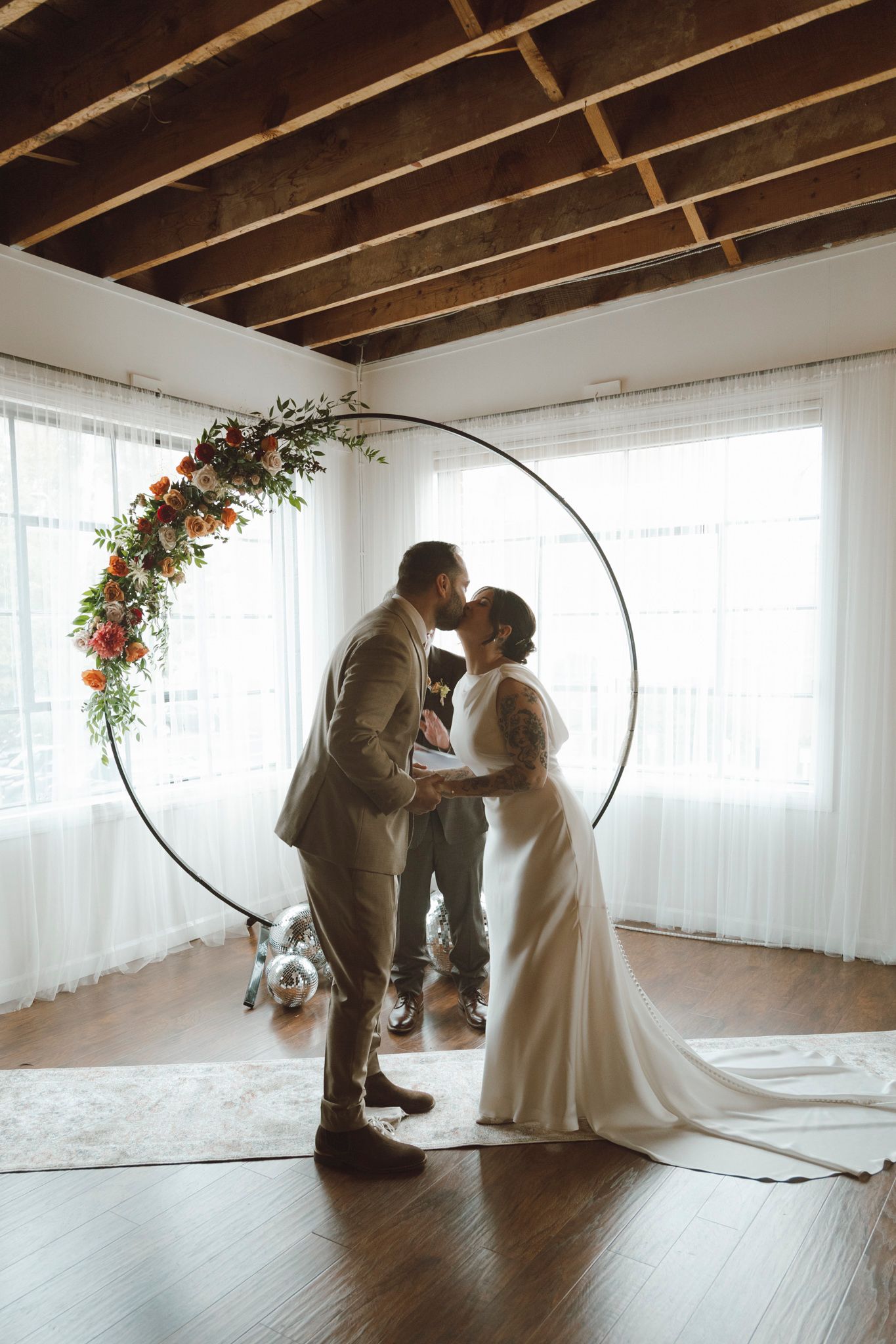 A couple shares an intimate dance moment in a bright sunlit room with exposed wooden beams and floral decorations.