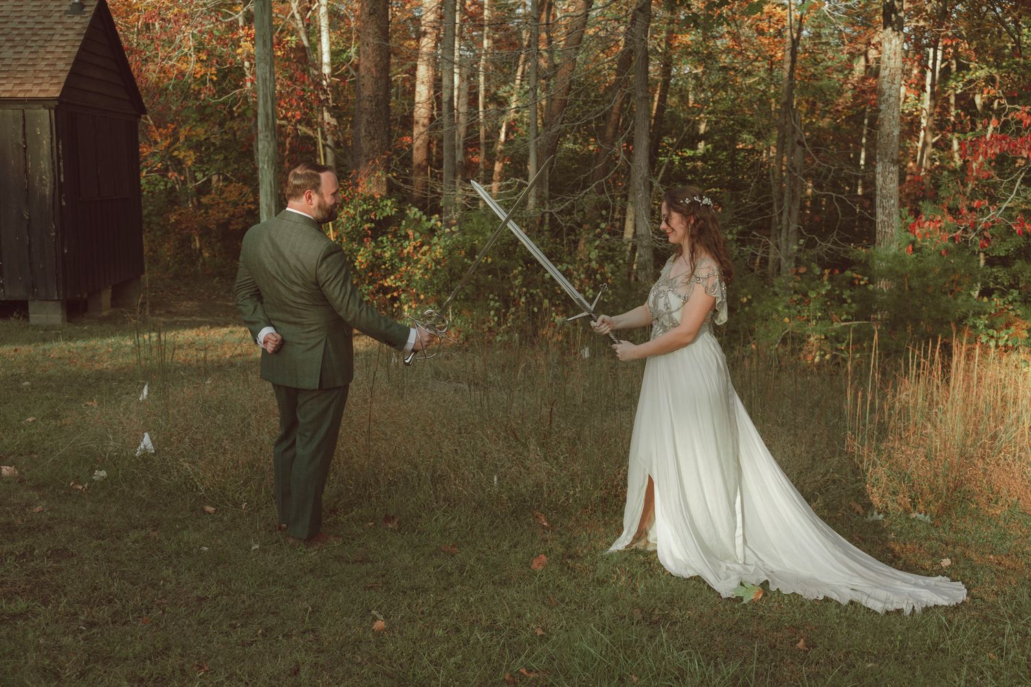 A couple in wedding attire playfully dueling with swords in an autumn forest setting.