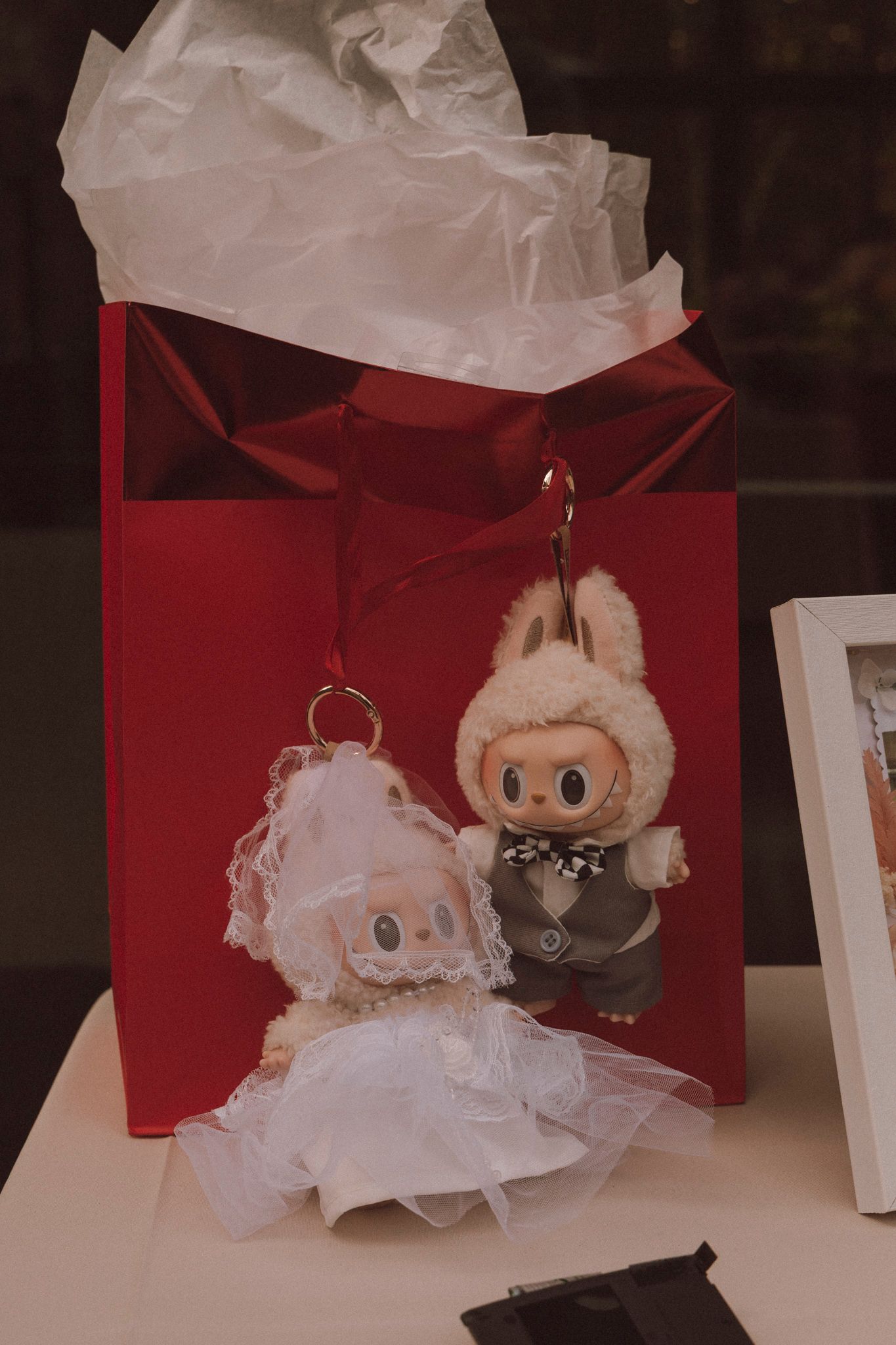 Two vintage kewpie doll ornaments with white and brown outfits sit beside a red gift bag with white tissue paper.