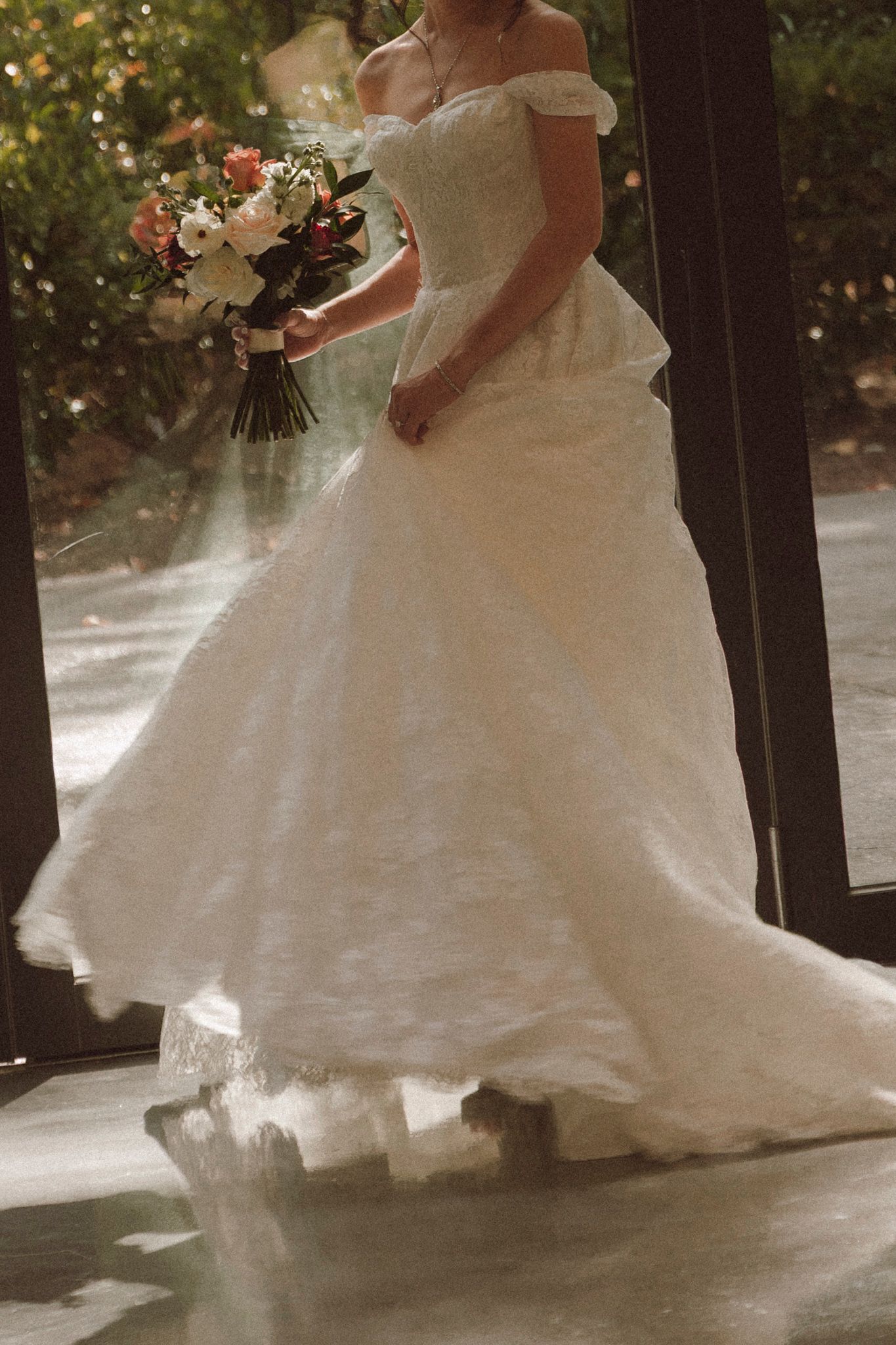 Elegant off-shoulder white wedding gown with flowing train and floral bouquet captured in dreamy backlit setting.