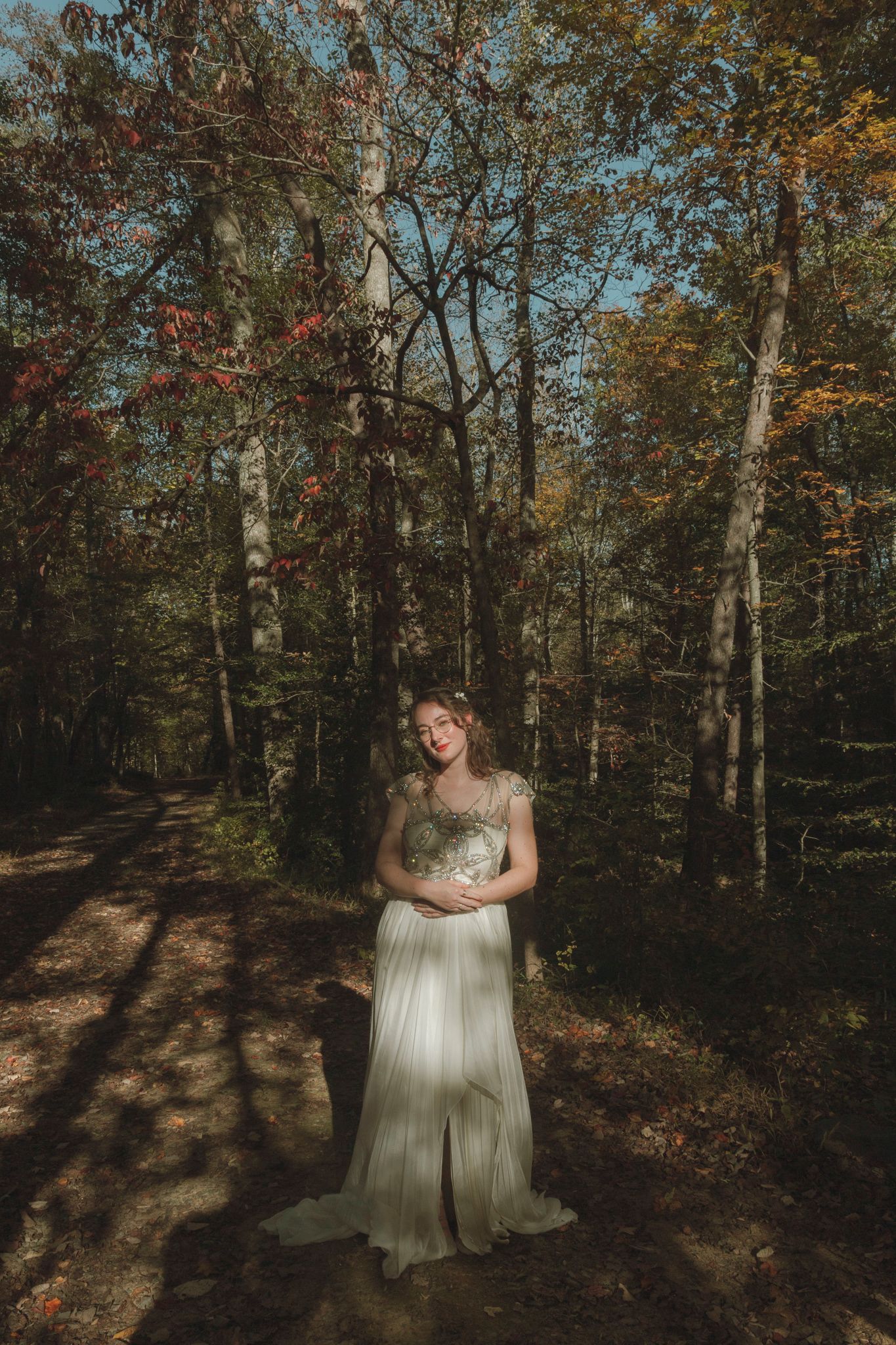 A figure in a flowing cream-colored dress stands in a sunlit autumn forest with golden leaves and tall trees.