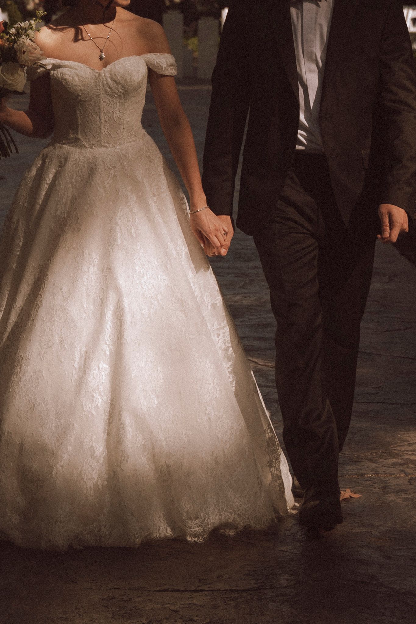 A couple holding hands at a wedding, with the bride wearing an off-shoulder ballgown and the groom in a black suit.