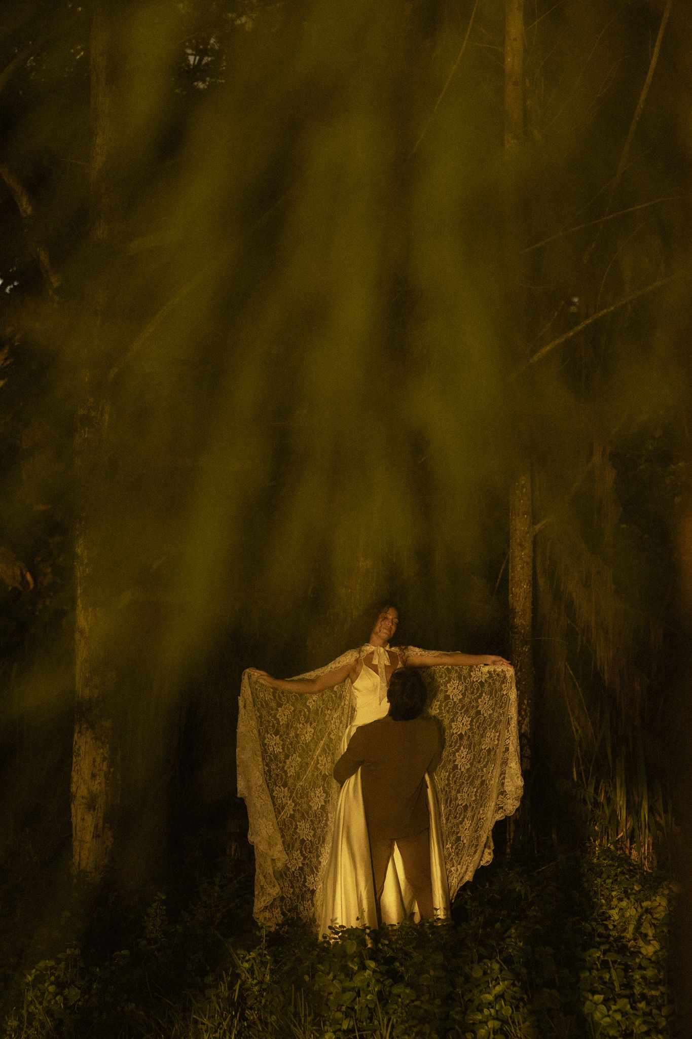 A woman in a white lace gown stands in a forest, bathed in golden light rays filtering through trees.