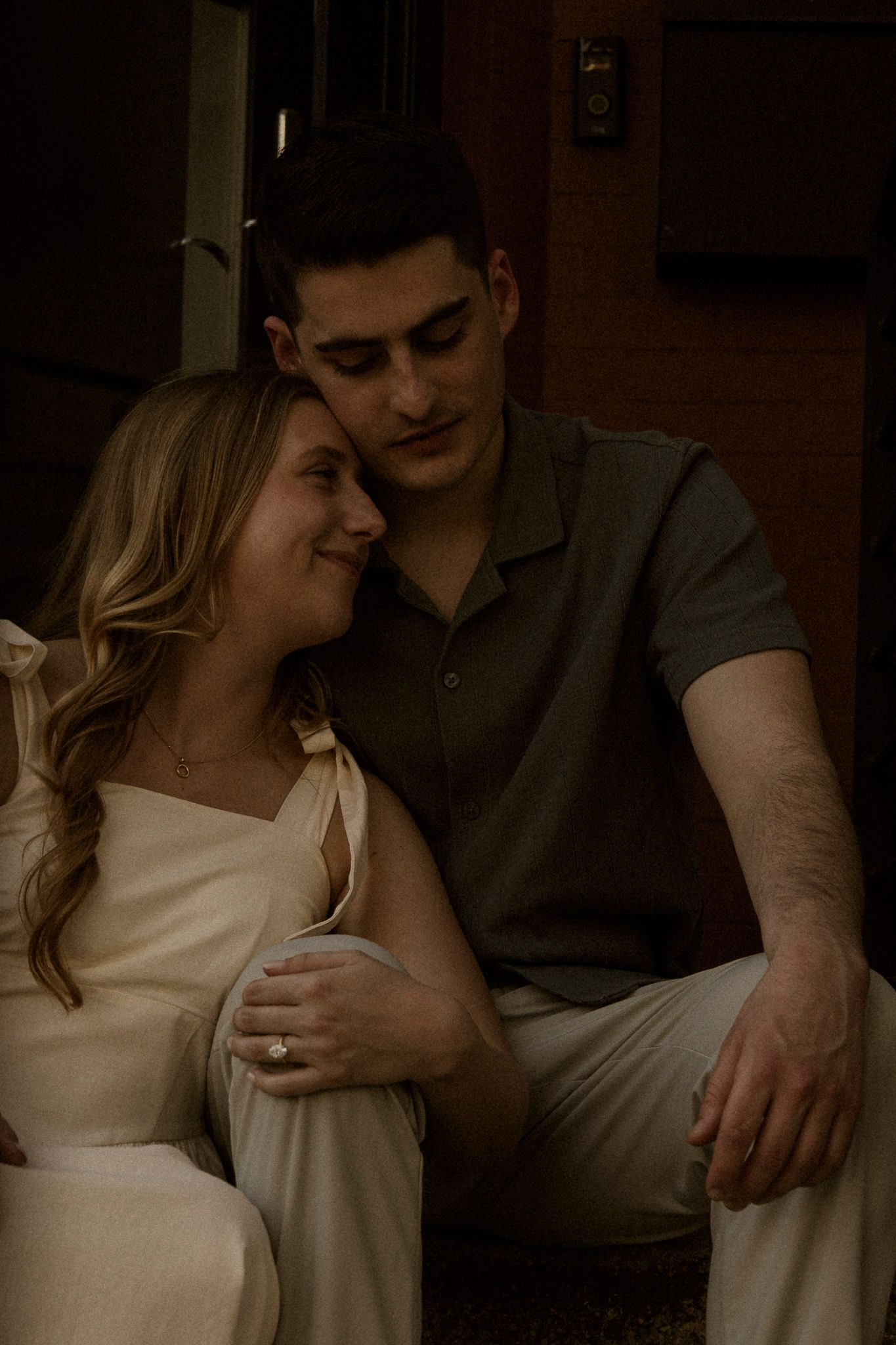 A couple sits closely together in warm light, woman in white dress smiling up at man in green polo shirt.