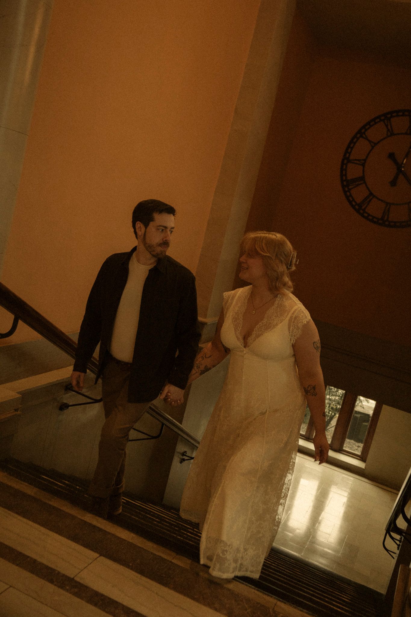 A couple stands together on stairs in a vintage building with warm lighting and an antique clock on the wall.