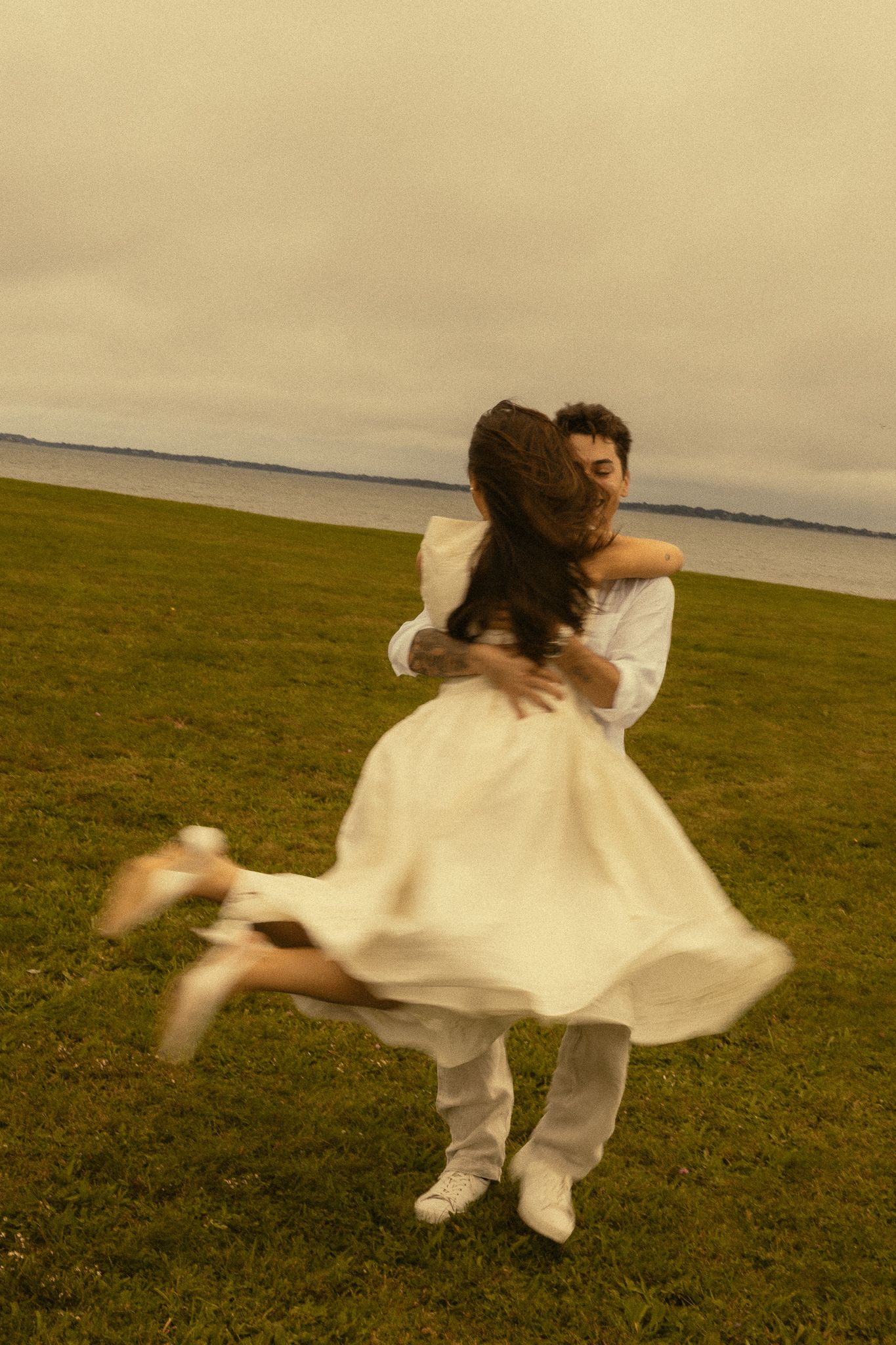 Couple in white clothing spinning joyfully on green grass near water under overcast sky.