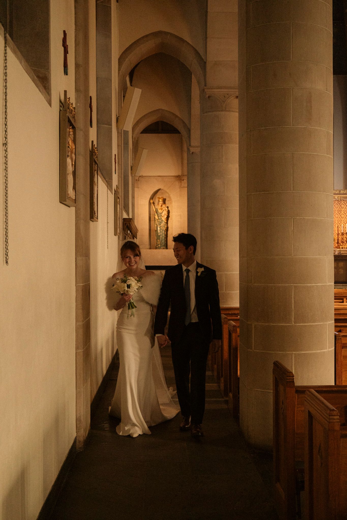 Bride and groom walking together through a warmly lit stone church aisle on their wedding day.