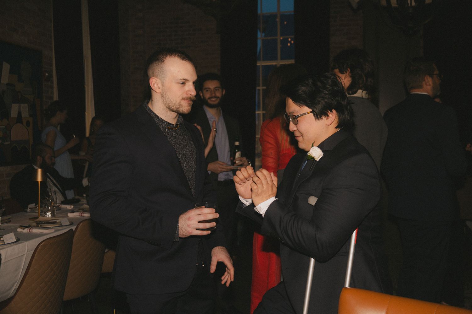 Two men in suits shaking hands at an elegant evening networking event with guests in background.