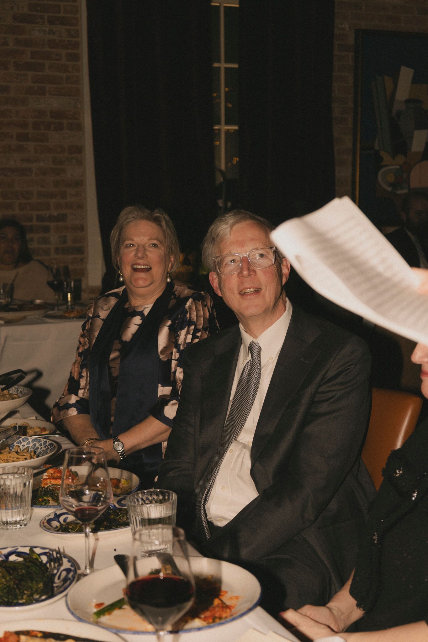 Elderly couple smiling at wedding reception dinner table with wine glasses and food.