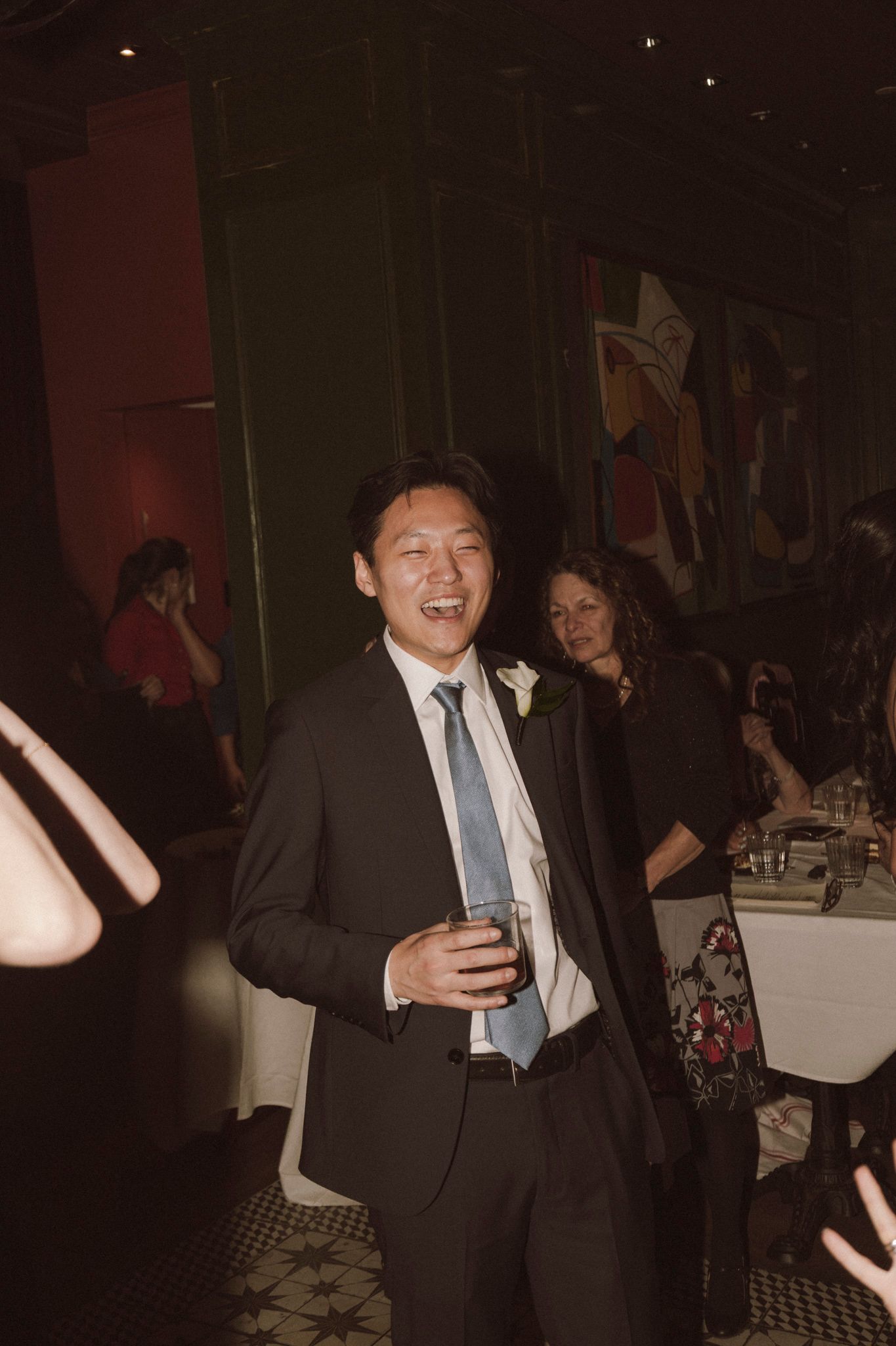 Smiling Asian man in dark suit and blue tie holding drink at an elegant indoor reception event.