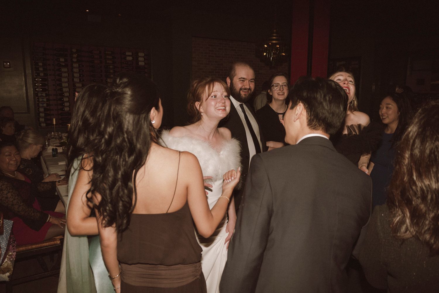 Bride in white feather wrap dancing and laughing with wedding guests on the reception dance floor.