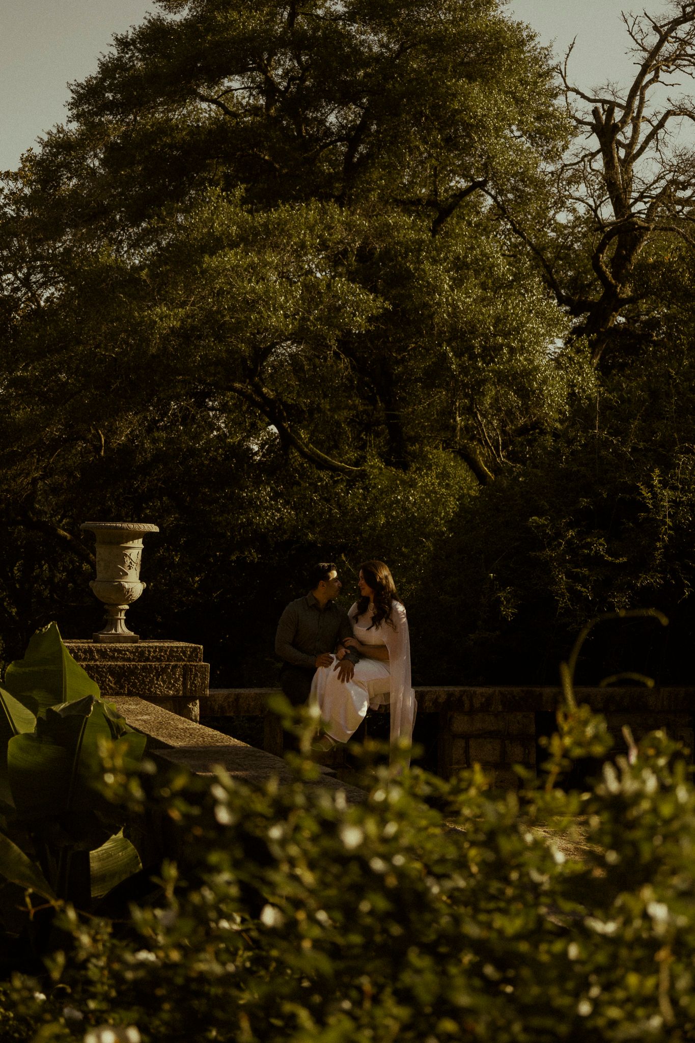 Couple in wedding attire sharing romantic moment in lush garden with stone urn and towering trees at golden hour.