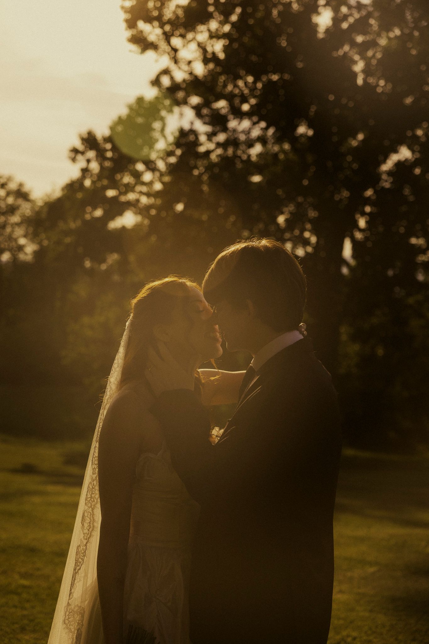Bride and groom share a romantic golden hour moment outdoors, bathed in warm sunset light with lush trees in the background.