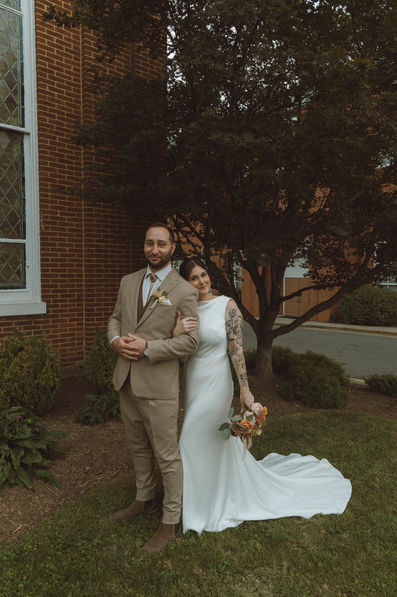 Couple in wedding attire poses outdoors against brick building with autumn foliage in background.